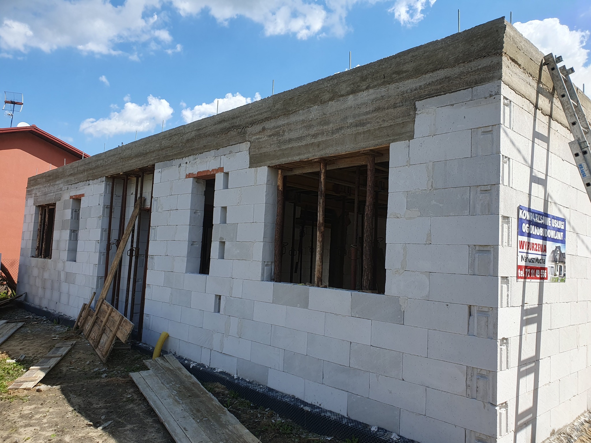 Ściana domu z betonu komórkowego w trakcie budowy, widoczne otwory okienne i wieniec. Na ścianie oparta drabina, w tle błękitne niebo.