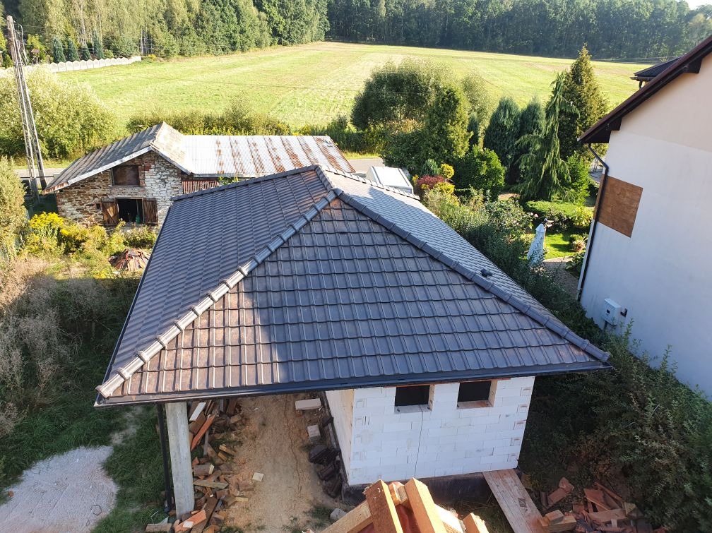 Nowy, grafitowy dach z dachówki ceramicznej na budynku w stanie surowym otwartym, z widokiem na wiejski krajobraz z zielonymi polami i drzewami.