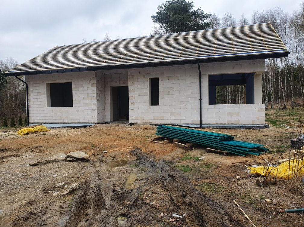 Surowy stan domu z białych bloczków, z widocznym dachem w trakcie budowy. Na pierwszym planie błoto i składowane deski, w tle las.