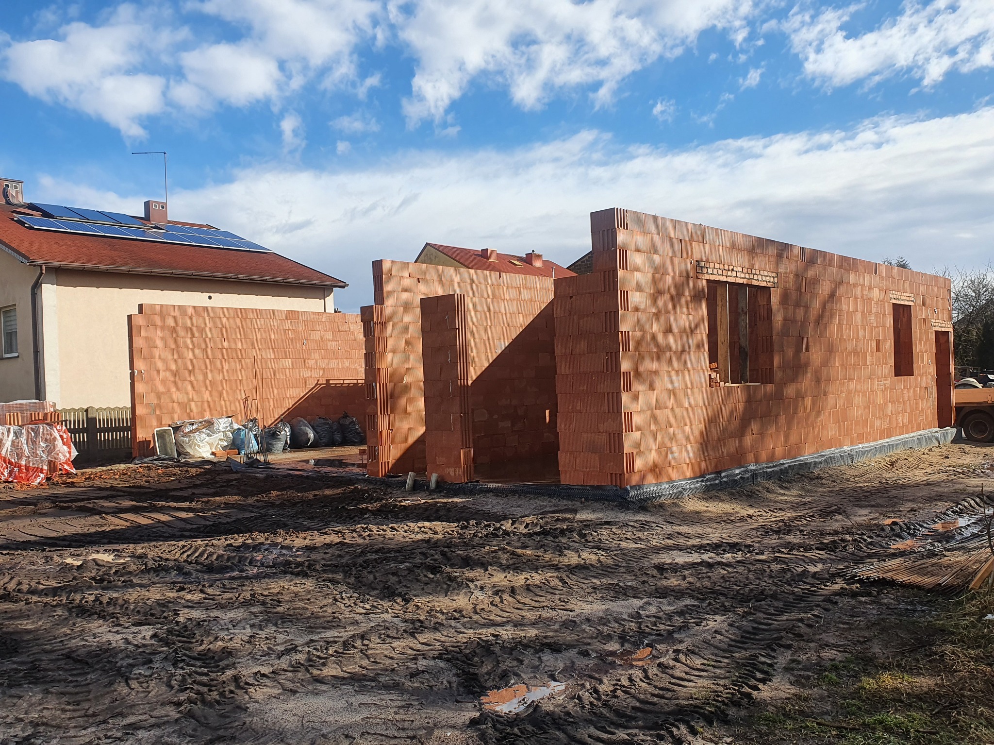 Wznoszenie ścian parterowego domu z bloczków ceramicznych. Widoczne otwory okienne i drzwiowe. Na dachu budynku obok panele słoneczne.