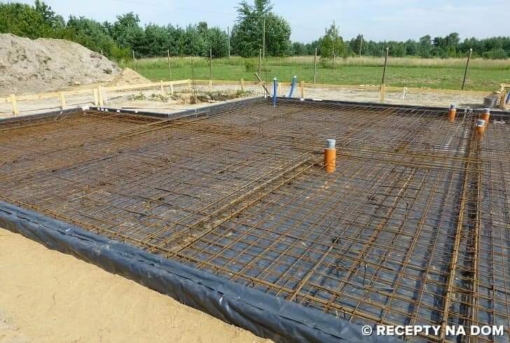 Zbrojenie fundamentów na czarnej folii, przygotowane do zalania betonem. Widoczne wystające rury instalacyjne i drewniane szalunki w tle. Dzień, teren budowy.