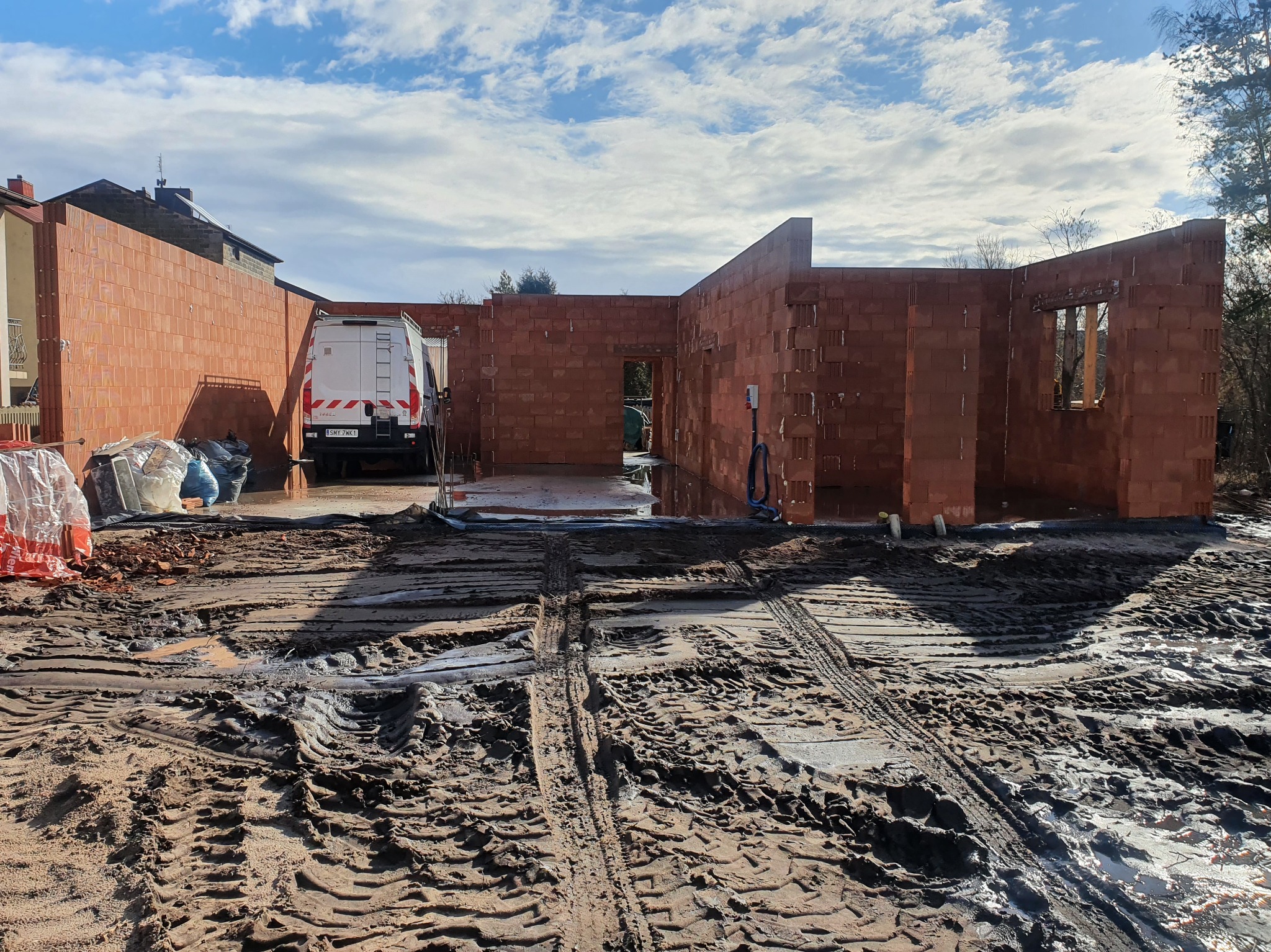 Budowa domu w trakcie wznoszenia ścian z czerwonej cegły. Na pierwszym planie błoto z wyraźnymi śladami opon. W tle widoczny biały samochód dostawczy.