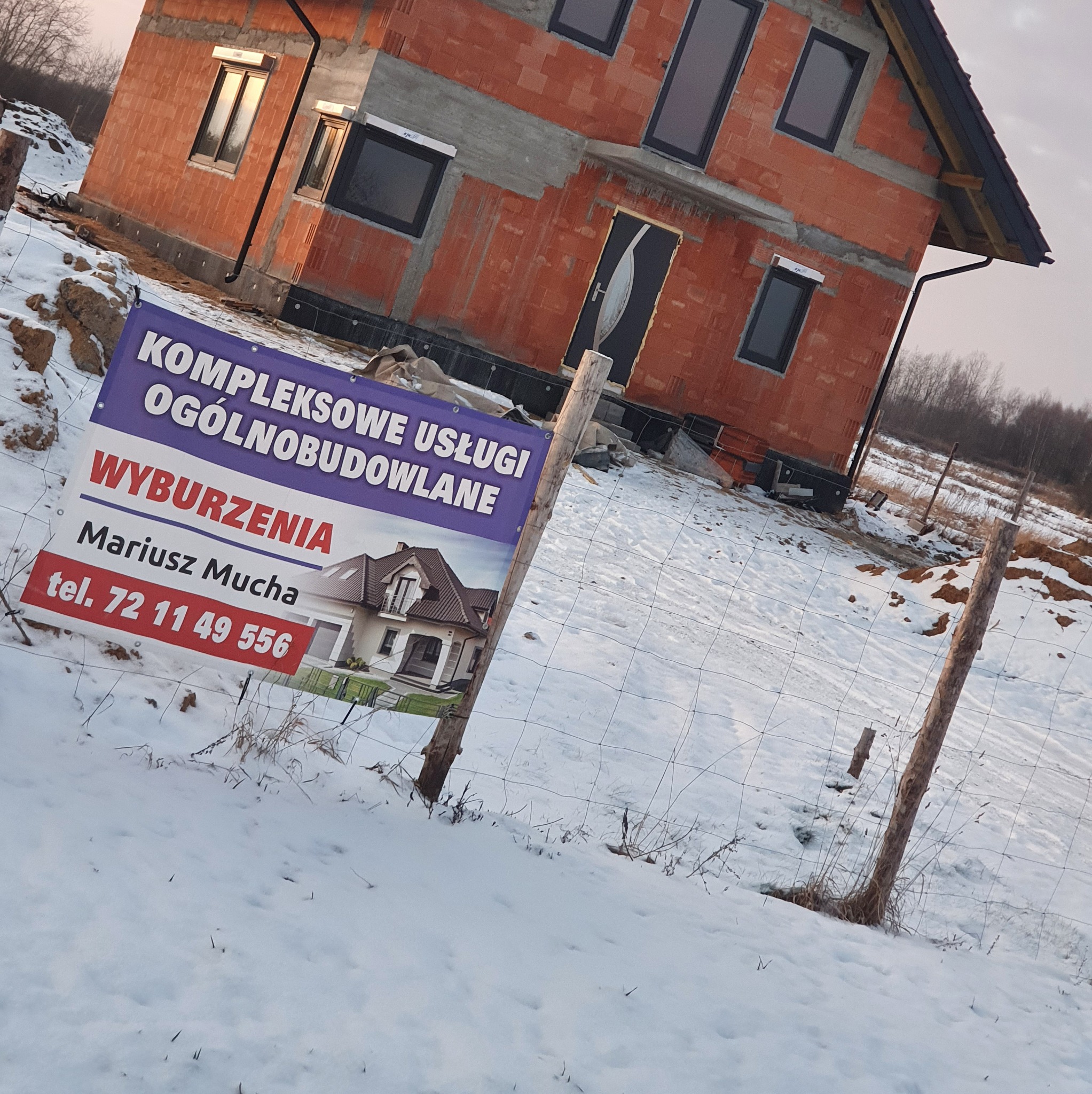 Budowa domu z czerwonej cegły w zimowym krajobrazie. Na pierwszym planie baner reklamowy z wizualizacją gotowego budynku i danymi kontaktowymi firmy.