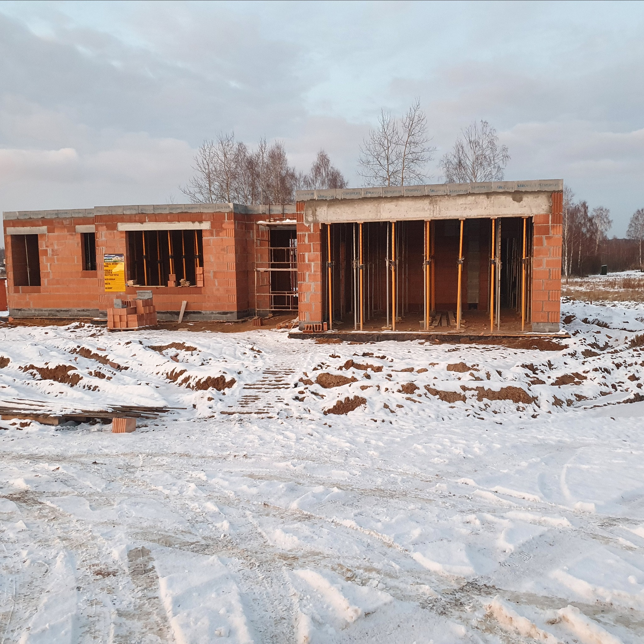 Budowa domu w stanie surowym zimą. Widoczne ściany z czerwonej cegły, drewniane podpory, śnieg i drzewa w tle. Częściowo ukończona konstrukcja.