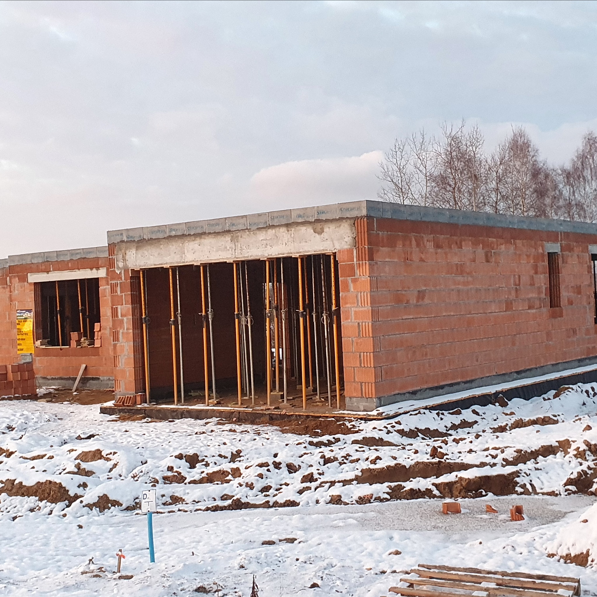 Surowy stan budynku z czerwonej cegły, zabezpieczony podporami. Zima, śnieg na ziemi. Widoczne drzewa w tle. Budowa w toku, bez wykończenia.