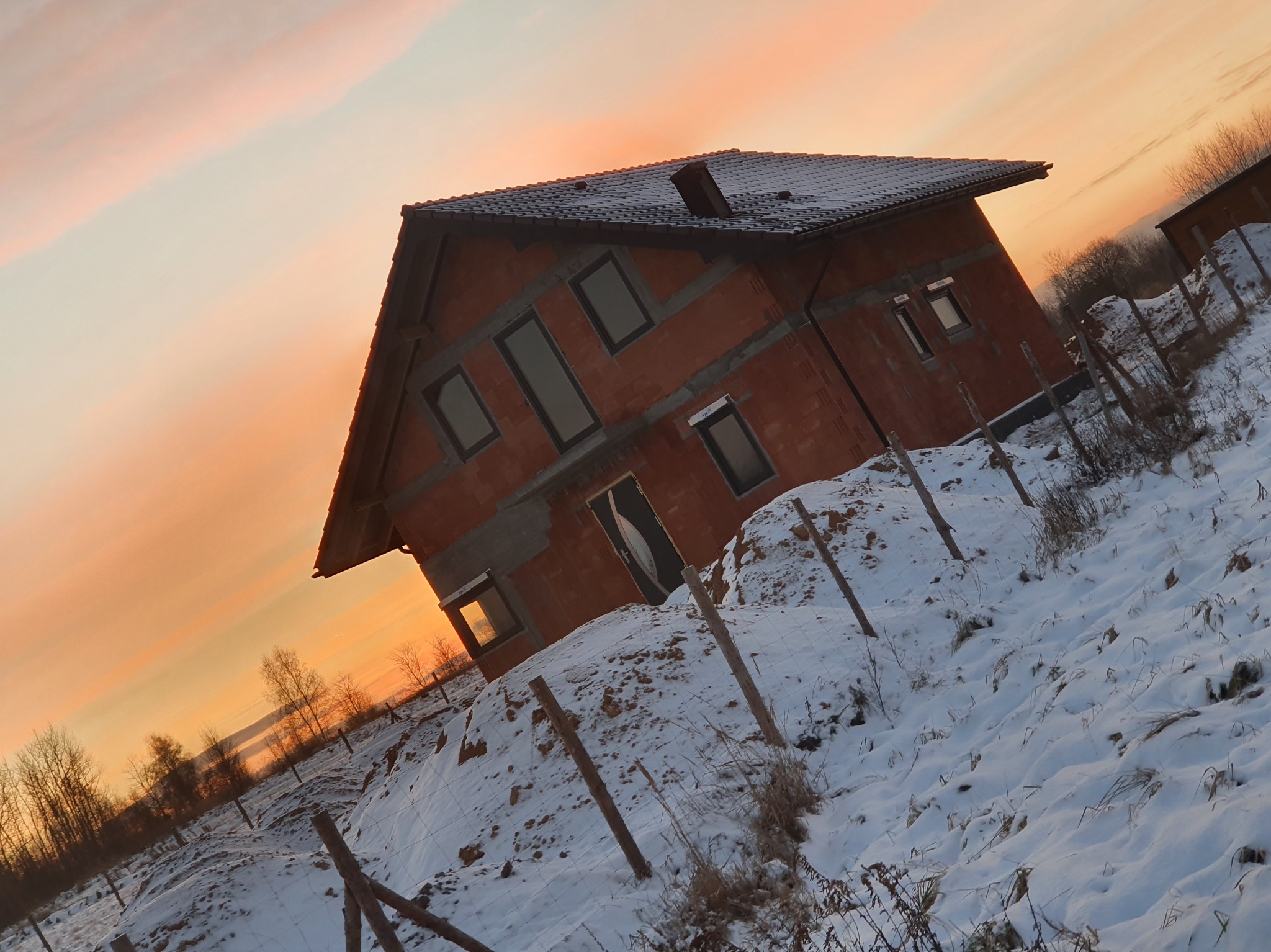 Surowy dom z cegły w zimowym krajobrazie o zachodzie słońca w Myszkowie. Widoczne okna, dach z dachówki i zasypane śniegiem otoczenie.