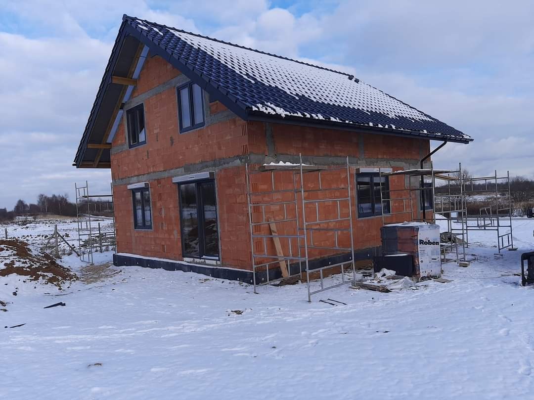 Surowy, dwukondygnacyjny dom z czerwonej cegły, z ciemną dachówką pokrytą śniegiem. Widoczne rusztowania i okna w czarnych ramach. Zimowy krajobraz.