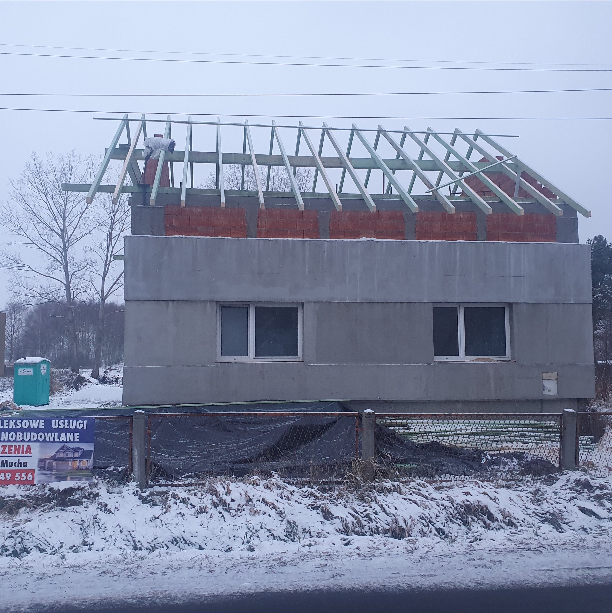 Budowa domu w zimowej scenerii: surowe ściany z pustaków, ceglana góra i drewniana konstrukcja dachu. Na pierwszym planie zasypany śniegiem teren.