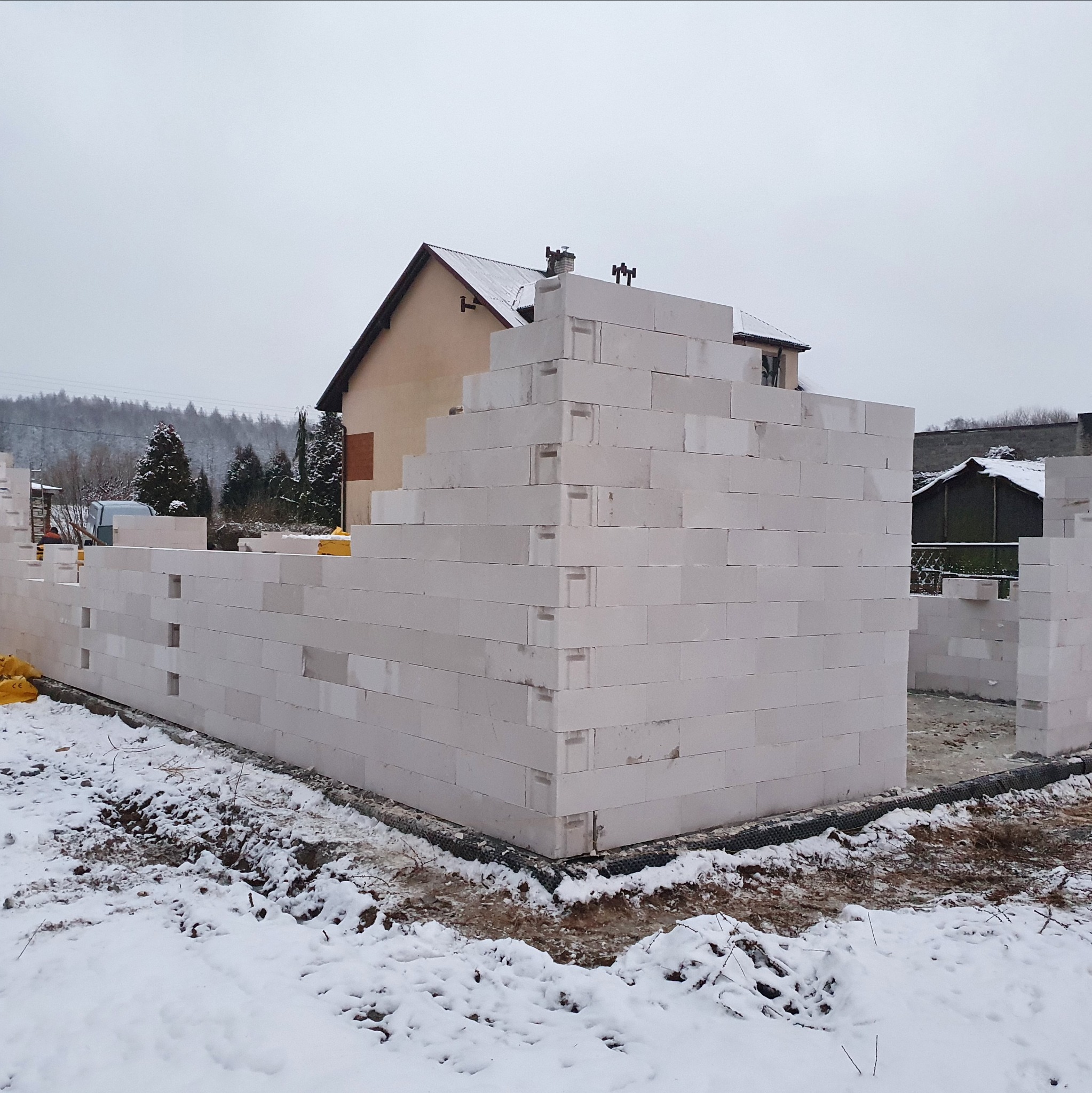 Ściany domu w trakcie murowania z białych bloczków, widoczne fundamenty i zimowy krajobraz. Częściowo wzniesione ściany.