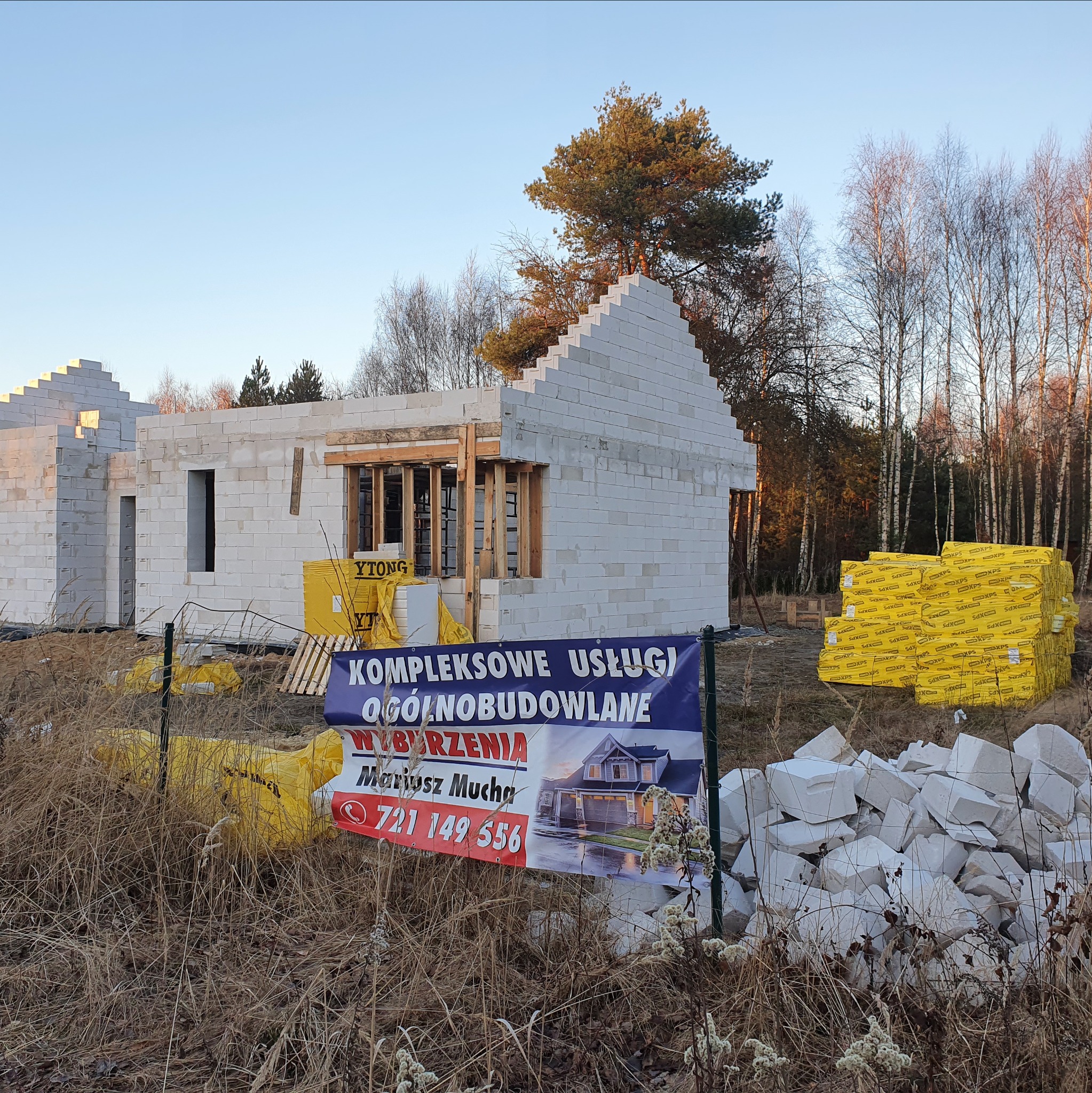 Wznoszenie ścian domu z bloczków Ytong. Na pierwszym planie reklama firmy budowlanej i stos materiałów izolacyjnych. Budowa w otoczeniu drzew.