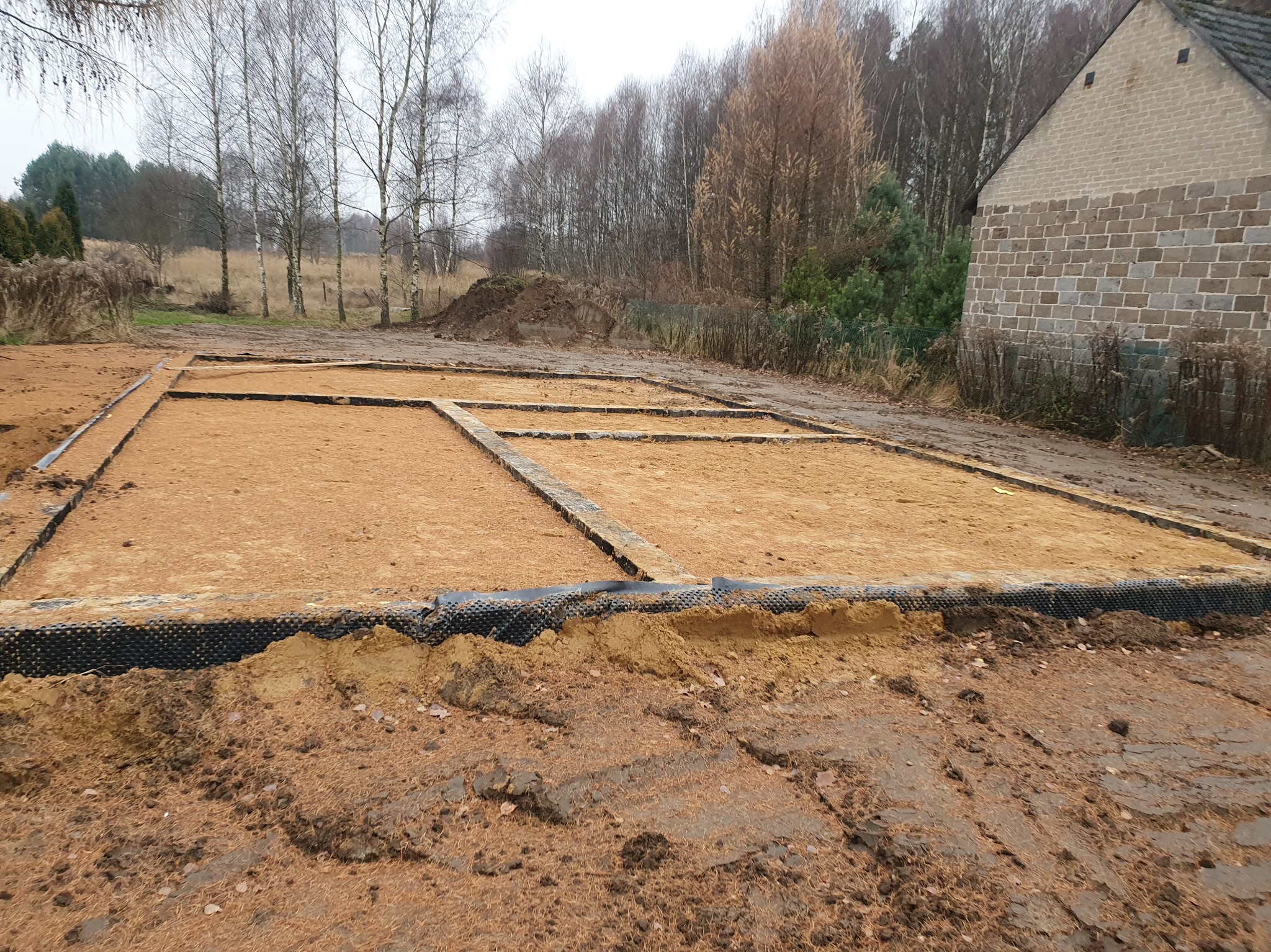 Przygotowany teren pod fundament domu z widoczną izolacją pionową i poziomą. W tle drzewa i fragment budynku z cegły.