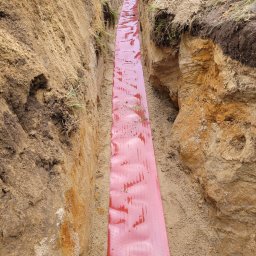 VoltBro - Wykop z ułożoną czerwoną folią ochronną kabla w ziemi. Ściany wykopu z piasku i ziemi. Wykop przygotowany pod ułożenie kabla.