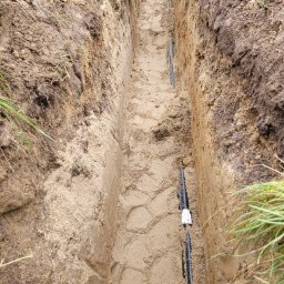 VoltBro - Wykop z ułożonymi rurami w piasku, prawdopodobnie pod kable, w Grzybnie. Widok z góry na wąski wykop z instalacją przed zasypaniem. Elementy instalacji elektrycznej.