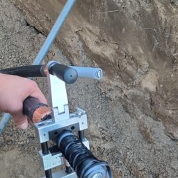 VoltBro - Podłączenie kabla elektrycznego w ziemi za pomocą specjalistycznego narzędzia. Widoczne przewody miedziane i warstwy izolacji. Wykop w ziemi z rurami w tle.