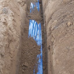 VoltBro - Wykop z ułożonymi niebieskimi rurami kablowymi, częściowo zasypanymi ziemią. Widok z góry na wąski wykop w ziemi.