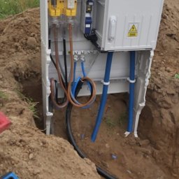 VoltBro - Szafa elektryczna z żółtymi elementami zabezpieczającymi, umieszczona w wykopie. Widoczne kable i poziomica.