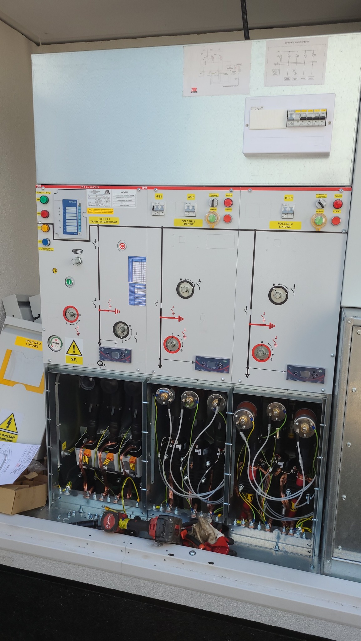 Rozdzielnica elektryczna z widocznymi elementami sterującymi, schematami i otwartymi komorami z okablowaniem w trakcie prac instalacyjnych. Narzędzia obok.