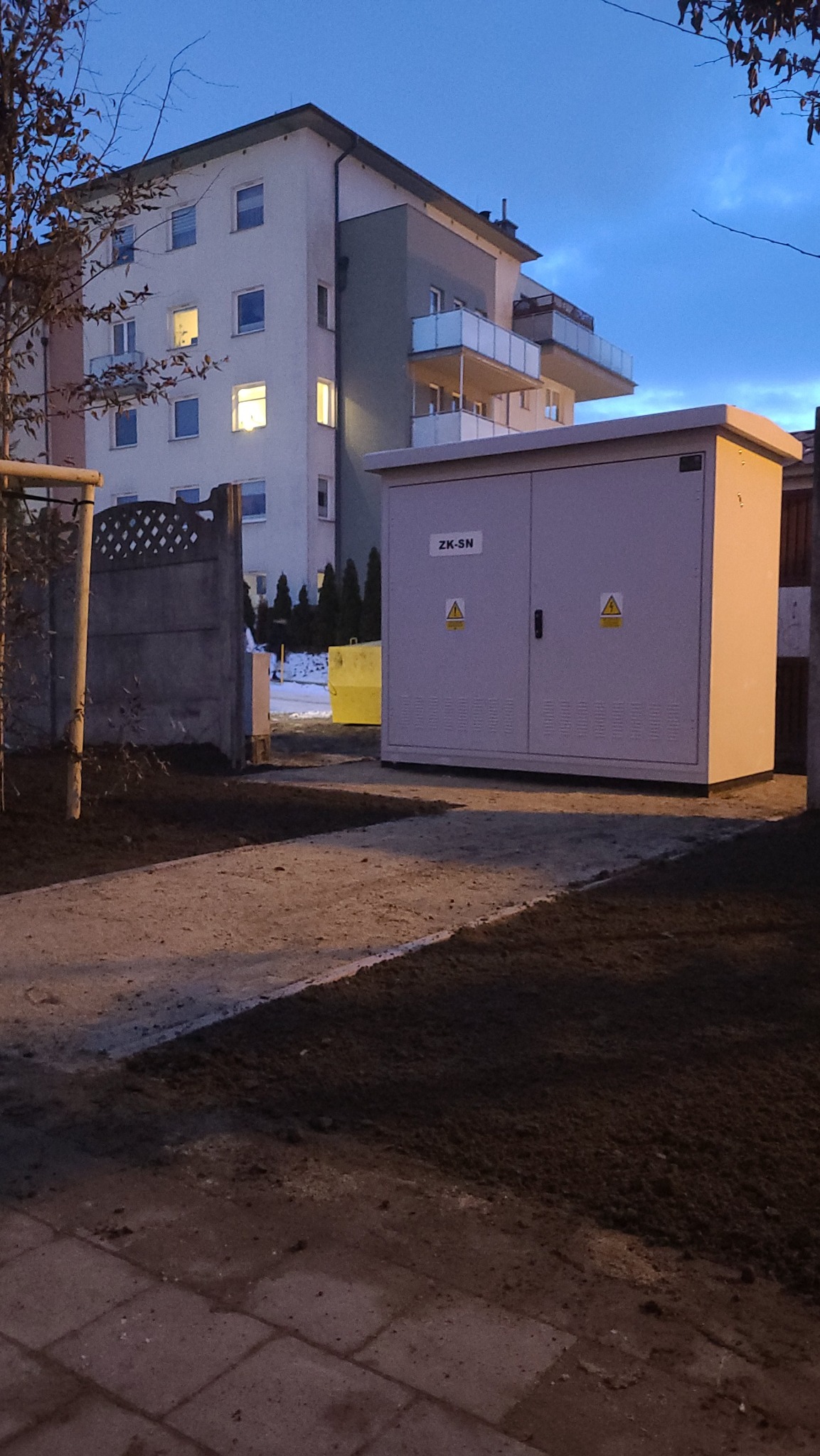 Zewnętrzna szafa elektryczna ZK-SN z ostrzeżeniami o napięciu na tle bloku mieszkalnego, sfotografowana o zmierzchu. Widoczny chodnik i świeżo obsiana ziemia.