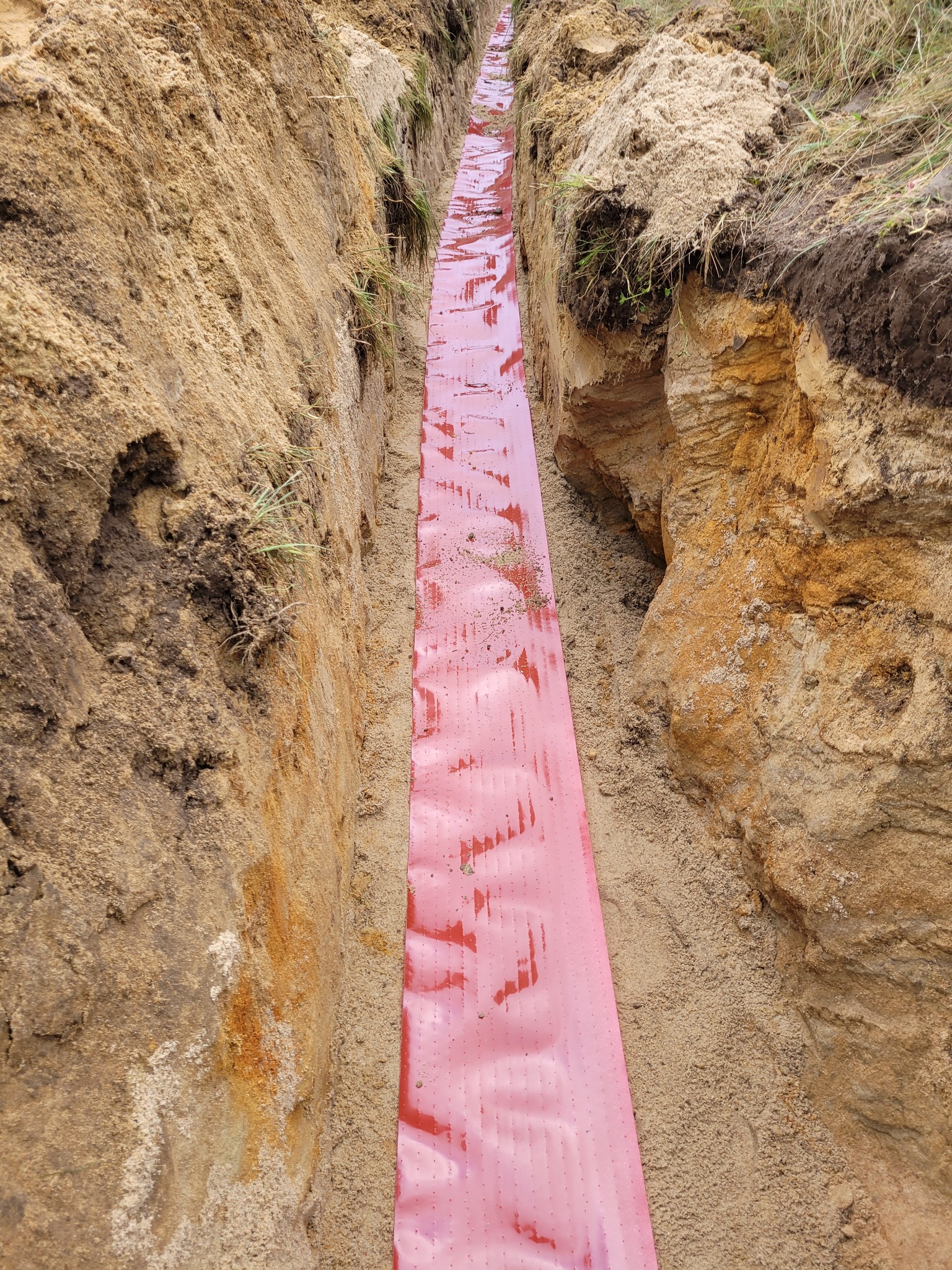 Wykop z ułożoną czerwoną folią ochronną kabla w ziemi. Ściany wykopu z piasku i ziemi. Wykop przygotowany pod ułożenie kabla.