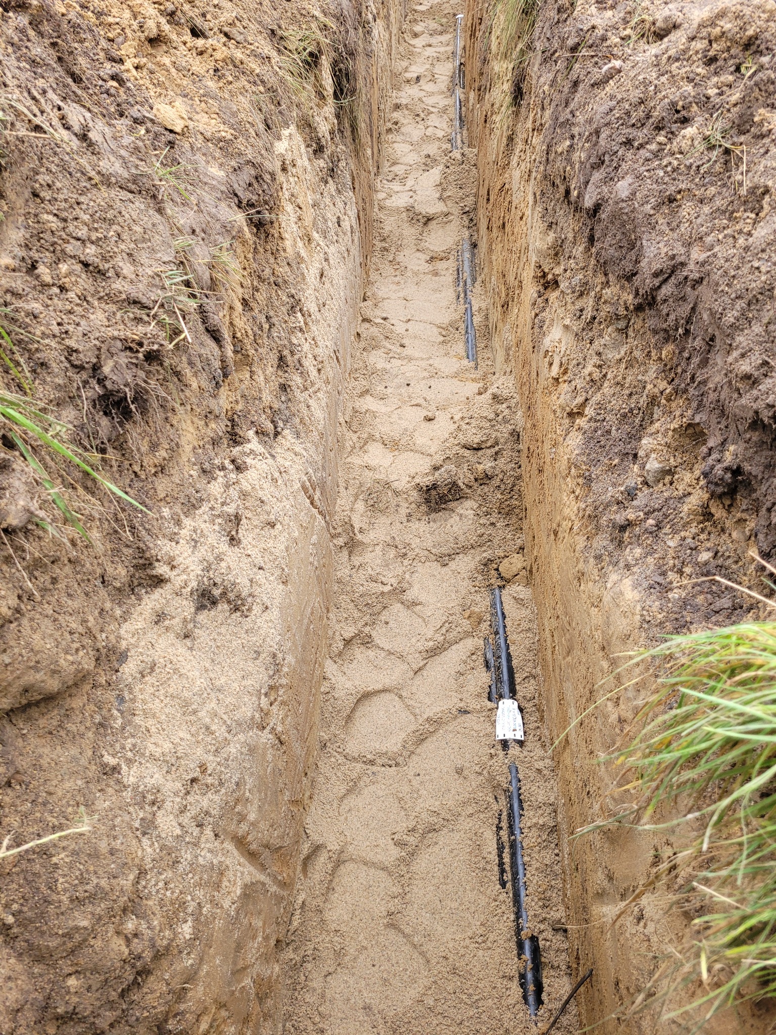Wykop z ułożonymi rurami w piasku, prawdopodobnie pod kable, w Grzybnie. Widok z góry na wąski wykop z instalacją przed zasypaniem. Elementy instalacji elektrycznej.