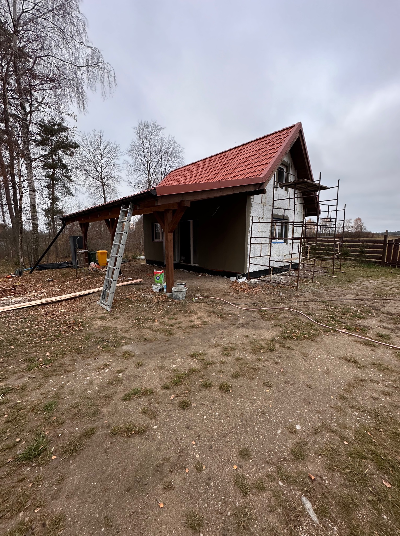 Niski domek w trakcie budowy: ocieplony styropianem, częściowo otynkowany, z czerwonym dachem. Widoczne rusztowania, drabina i materiały budowlane.