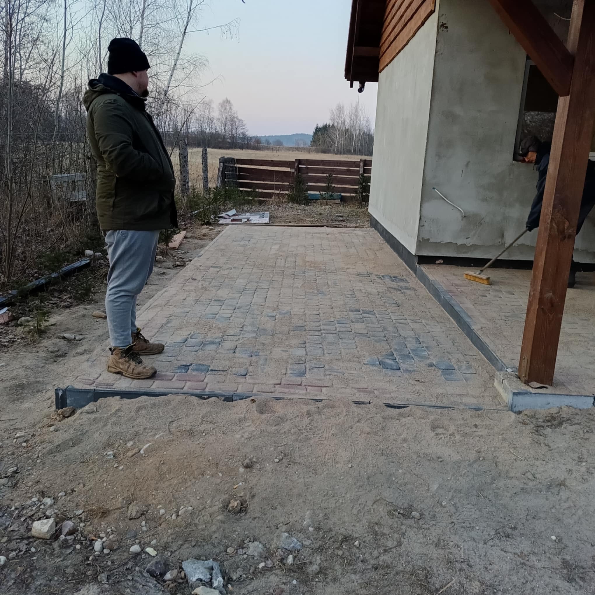 Układanie kostki brukowej na posesji przy domu. Widoczny pracownik zamiatający piasek. W tle drzewa i pole. Kąt ujęcia z dołu.
