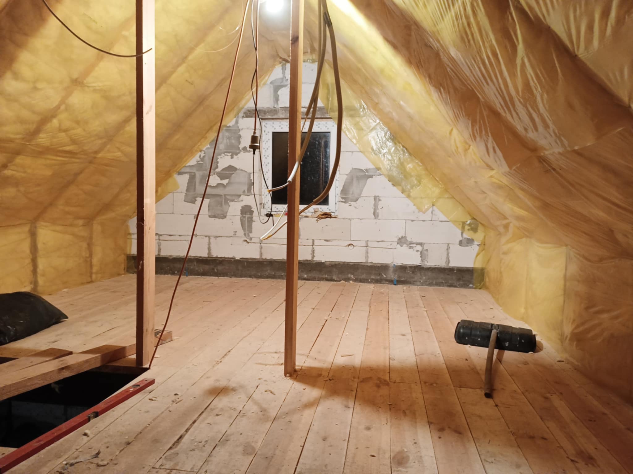 Adaptacja poddasza: drewniana podłoga, ocieplenie folią, ściany z bloczków. Widoczne kable i narzędzia. Prace wykończeniowe na strychu.