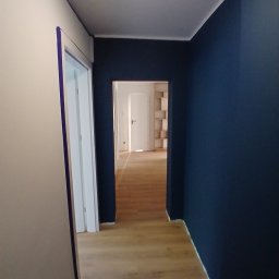 RAFAŁ CYLKOWSKI - Korytarz po malowaniu: ściana w odcieniu granatu kontrastuje z jasną podłogą i białymi ścianami. Widoczne taśmy malarskie przy drzwiach.