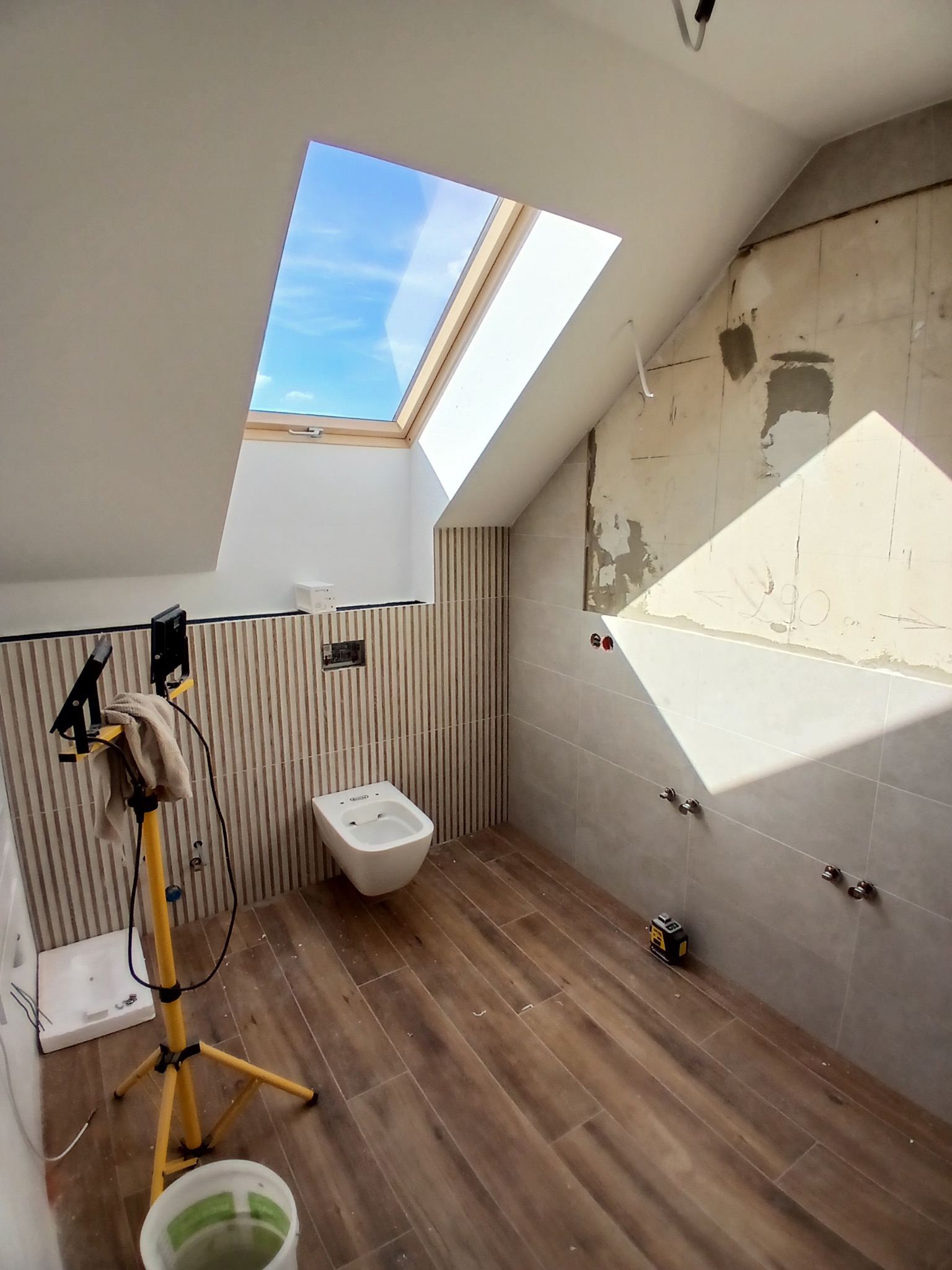 Ukończona łazienka na poddaszu z drewnopodobną podłogą, częściowo wykończona płytkami, z oknem dachowym i bidetem. Widoczny laser krzyżowy i lampa budowlana.