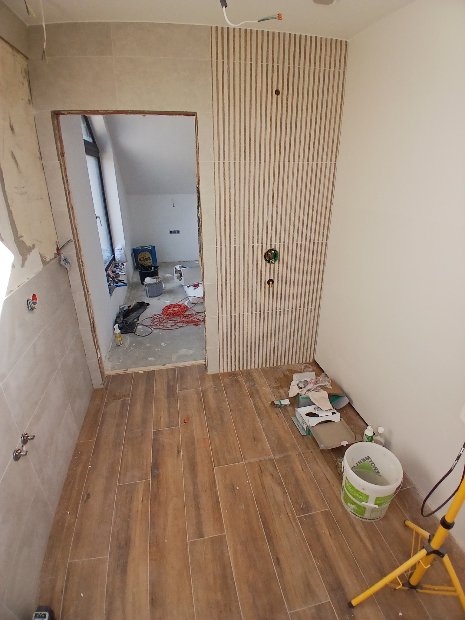 Remont łazienki w trakcie: ułożona podłoga z płytek imitujących drewno, ściana z dekoracyjnymi panelami pionowymi. Widoczne narzędzia i materiały budowlane.