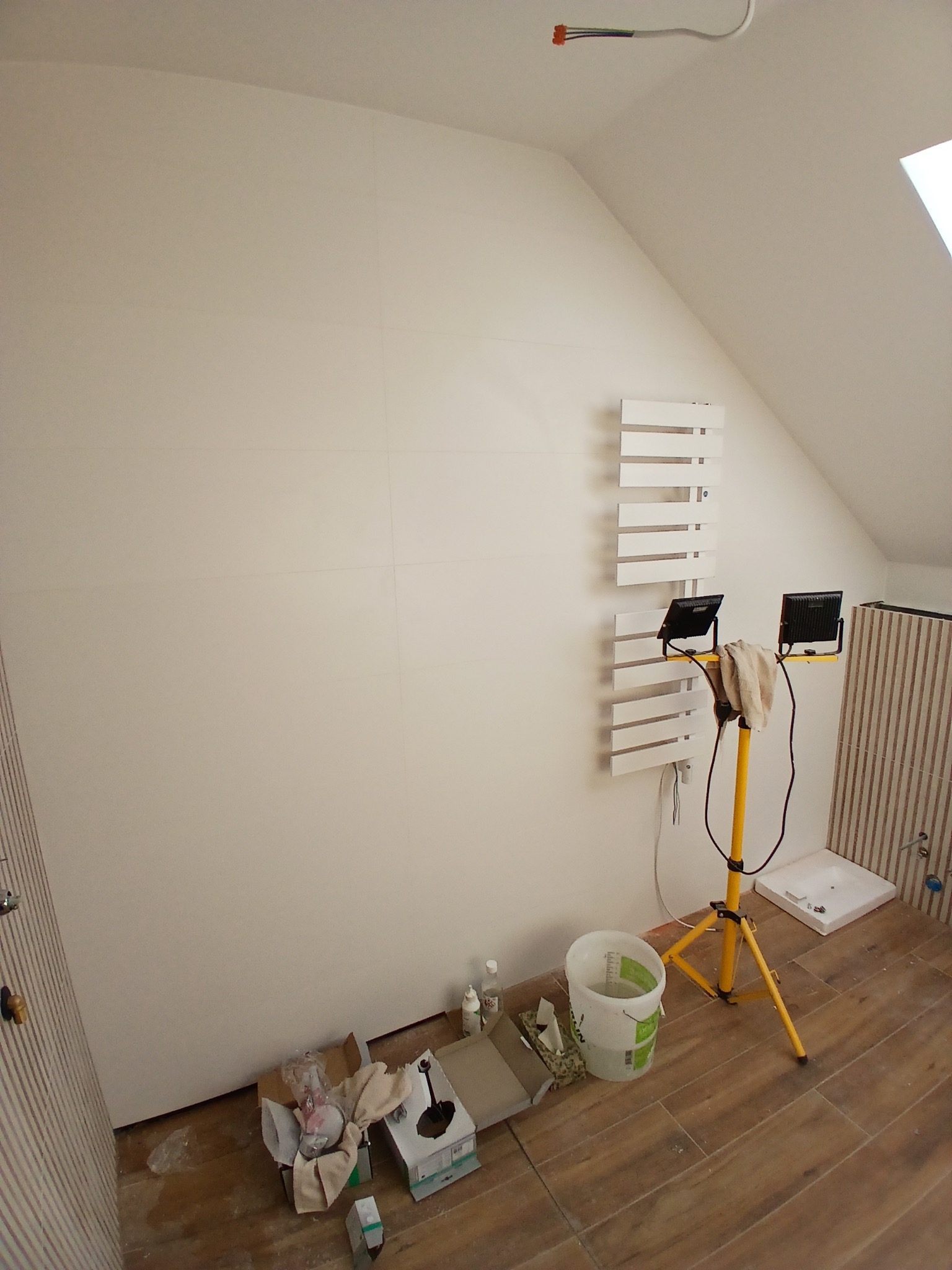 Biała ściana z grzejnikiem drabinkowym w Pęgowie, oświetlona lampą budowlaną. Widoczne narzędzia i materiały przygotowane do prac wykończeniowych w łazience na poddaszu.