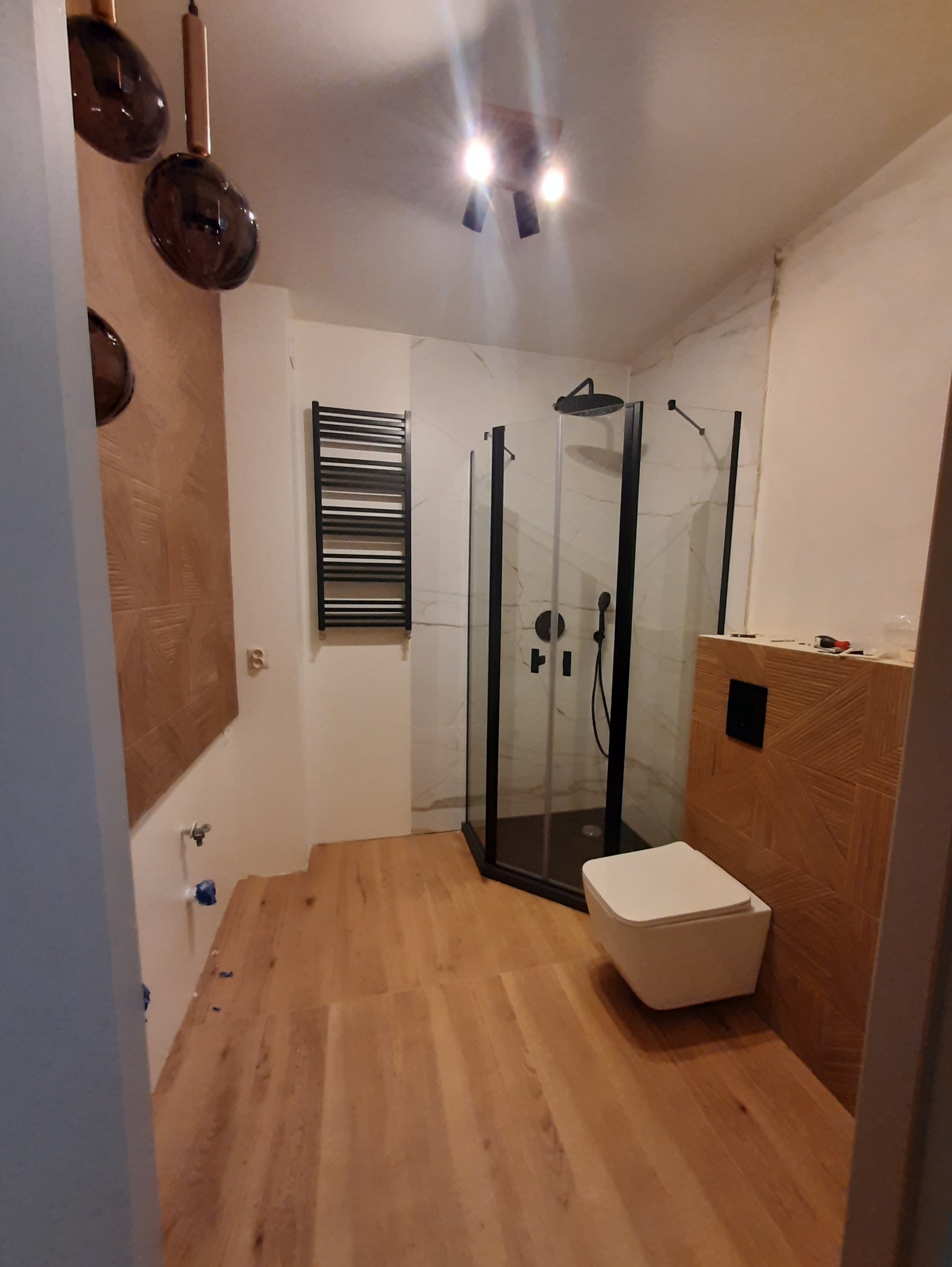 Nowoczesna łazienka z kabiną prysznicową typu walk-in, drewnianą podłogą i dekoracyjną ścianą. Czarny grzejnik drabinkowy i minimalistyczna toaleta podwieszana.