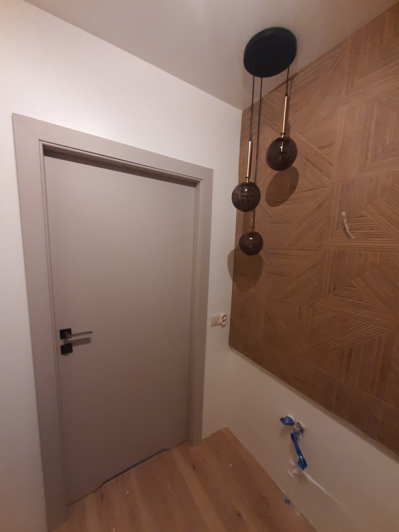 Beżowe drzwi z czarną klamką, drewniana ściana z geometrycznym wzorem i wisząca lampa z trzema szklanymi kulami w wykończonym wnętrzu.