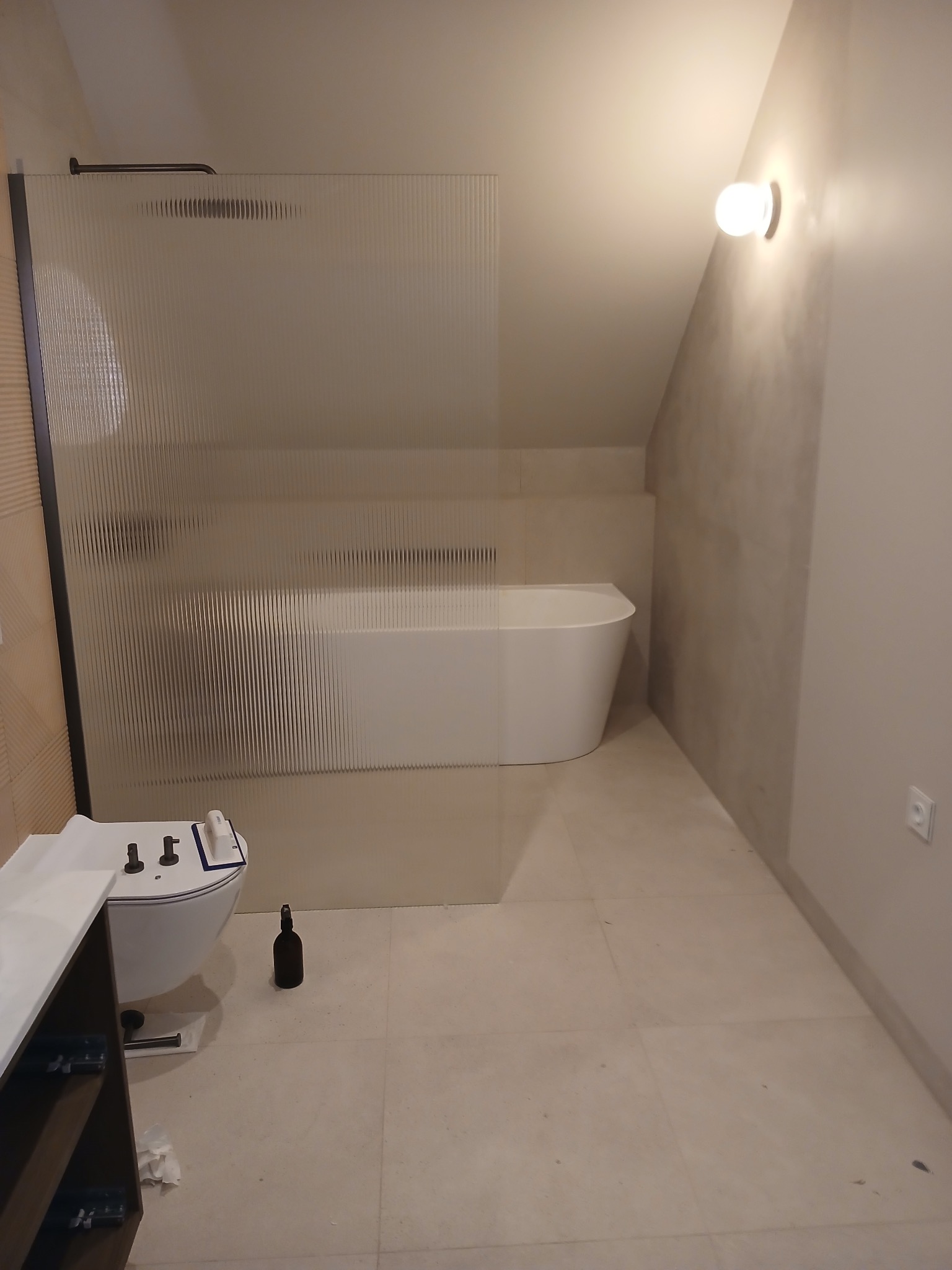 Nowoczesna łazienka w odcieniach beżu z kabiną prysznicową z ryflowanego szkła, wolnostojącą wanną i podwieszaną toaletą. Minimalistyczny design, oświetlenie punktowe.