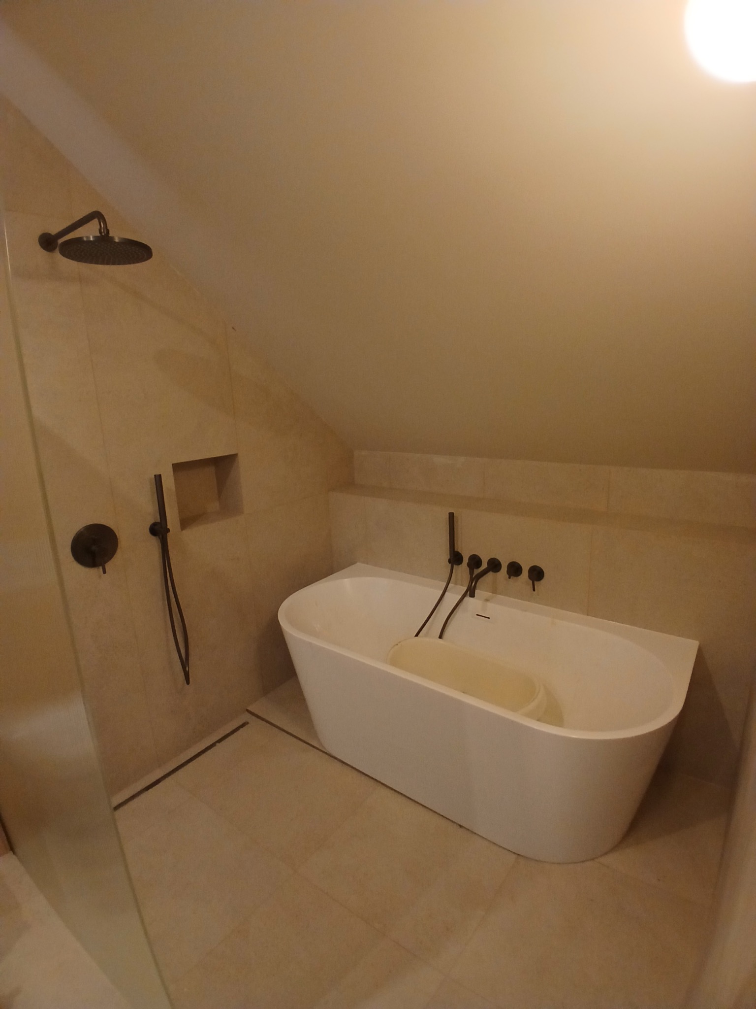 Nowoczesna łazienka na poddaszu z wolnostojącą białą wanną i czarną armaturą. Beżowe płytki na ścianach i podłodze. Ujęcie z kabiny prysznicowej.