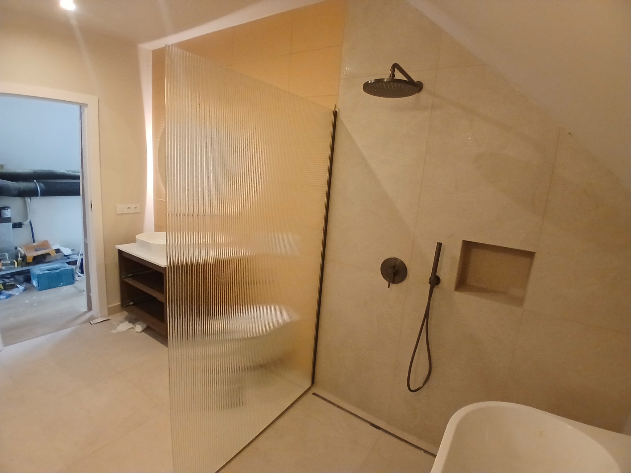 Nowoczesna łazienka z kabiną prysznicową oddzieloną ryflowaną szybą, armatura w kolorze brązu, beżowe płytki. W tle widoczny fragment szafki z umywalką i otwarte drzwi.
