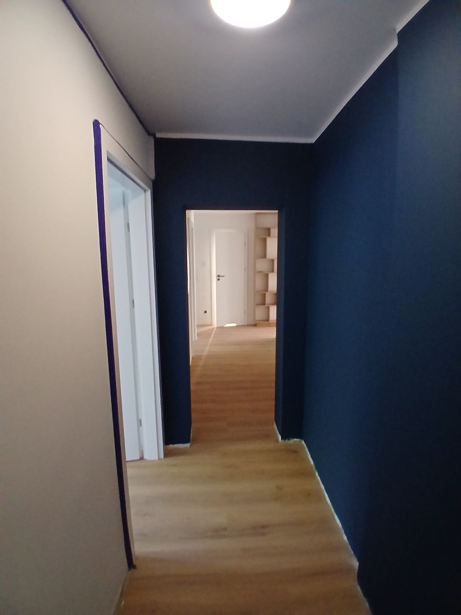 Korytarz po malowaniu: ściana w odcieniu granatu kontrastuje z jasną podłogą i białymi ścianami. Widoczne taśmy malarskie przy drzwiach.