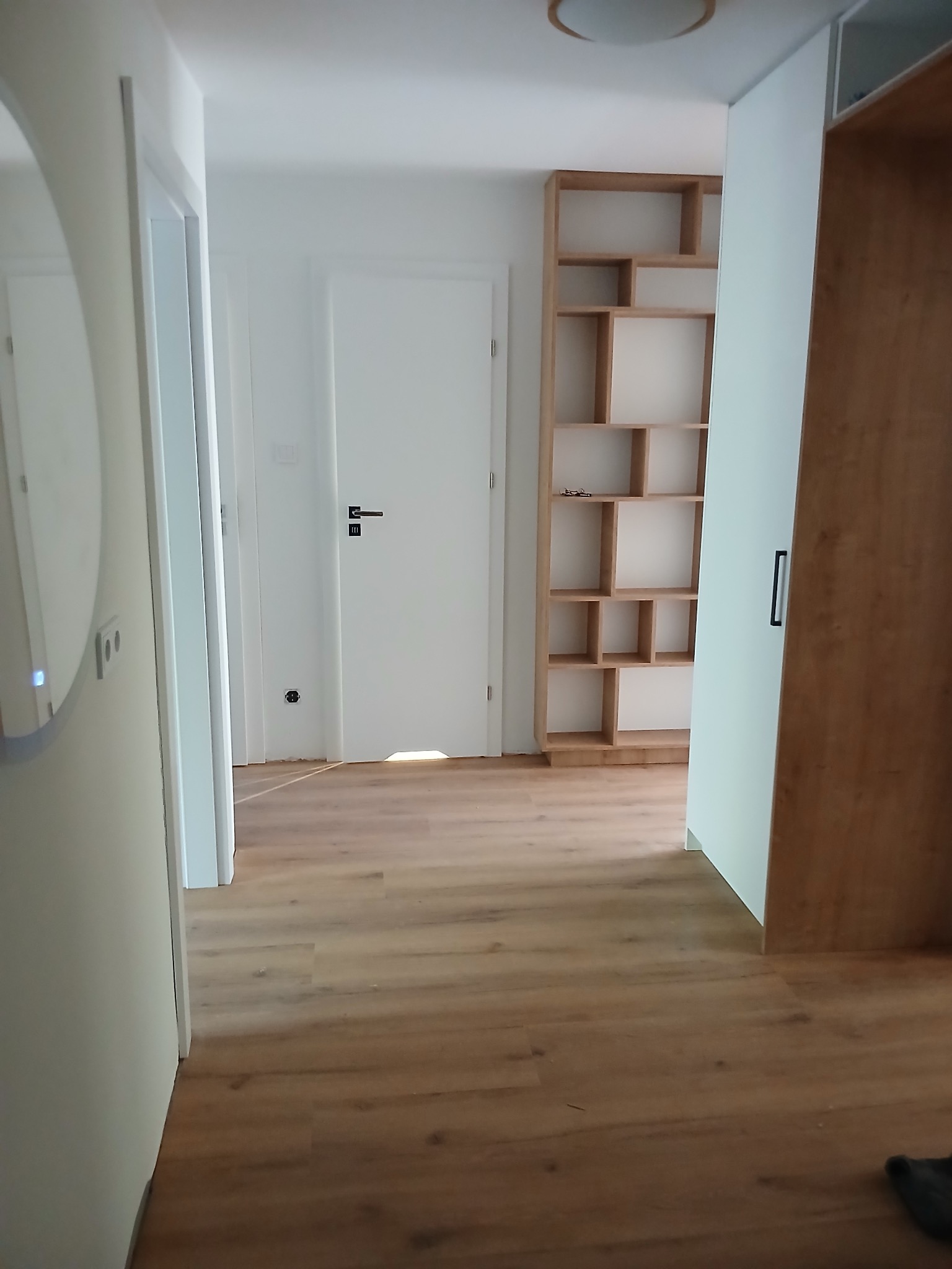 Jasne wnętrze po remoncie: drewniana podłoga, białe ściany i drzwi, regał z jasnego drewna. Minimalistyczny design, czyste linie, nowoczesne oświetlenie sufitowe.