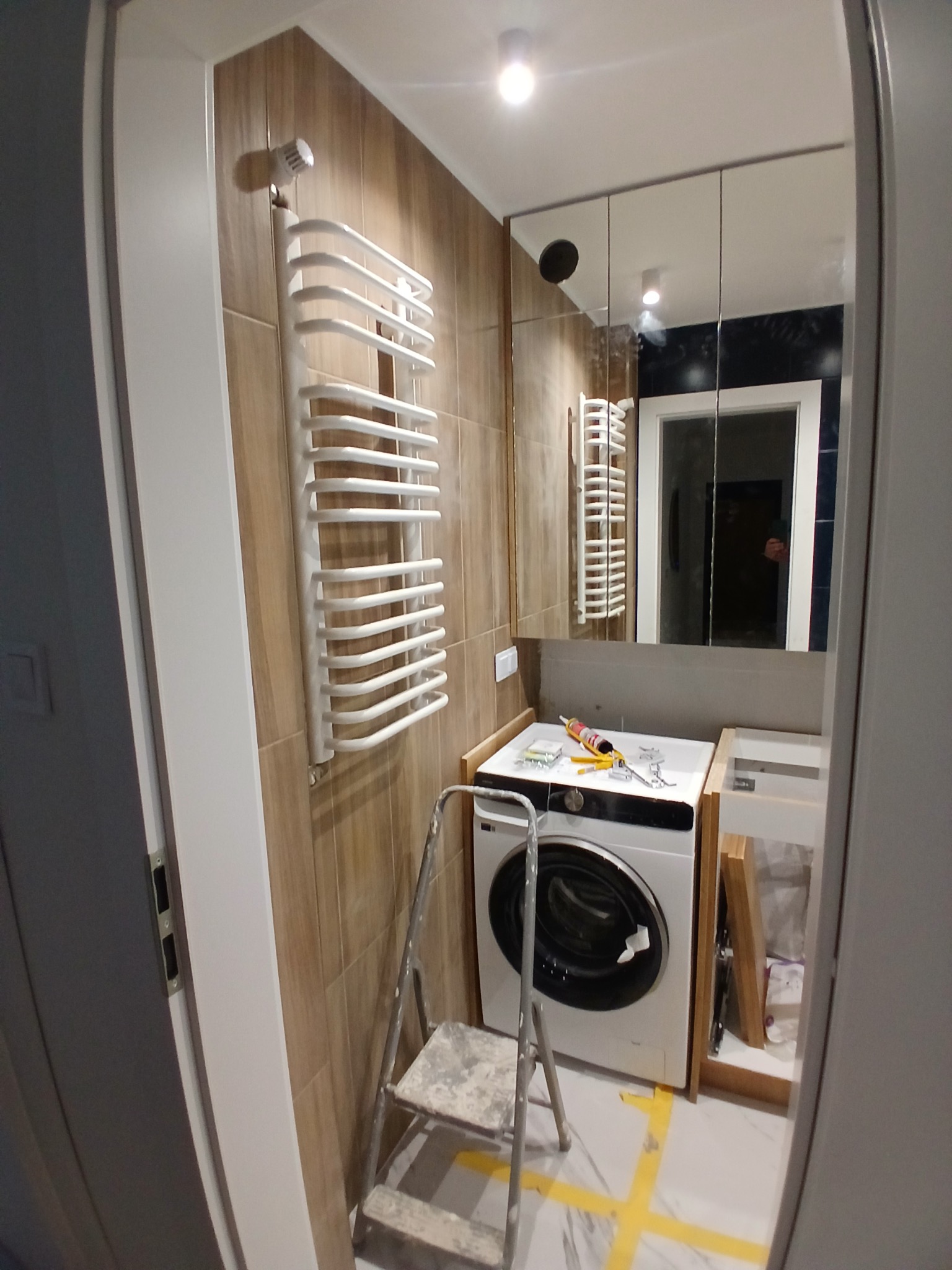 Łazienka w trakcie wykończenia: grzejnik drabinkowy na brązowych płytkach, pralka z silikonem, drabina malarska, lustro. Widoczne przygotowania do montażu.