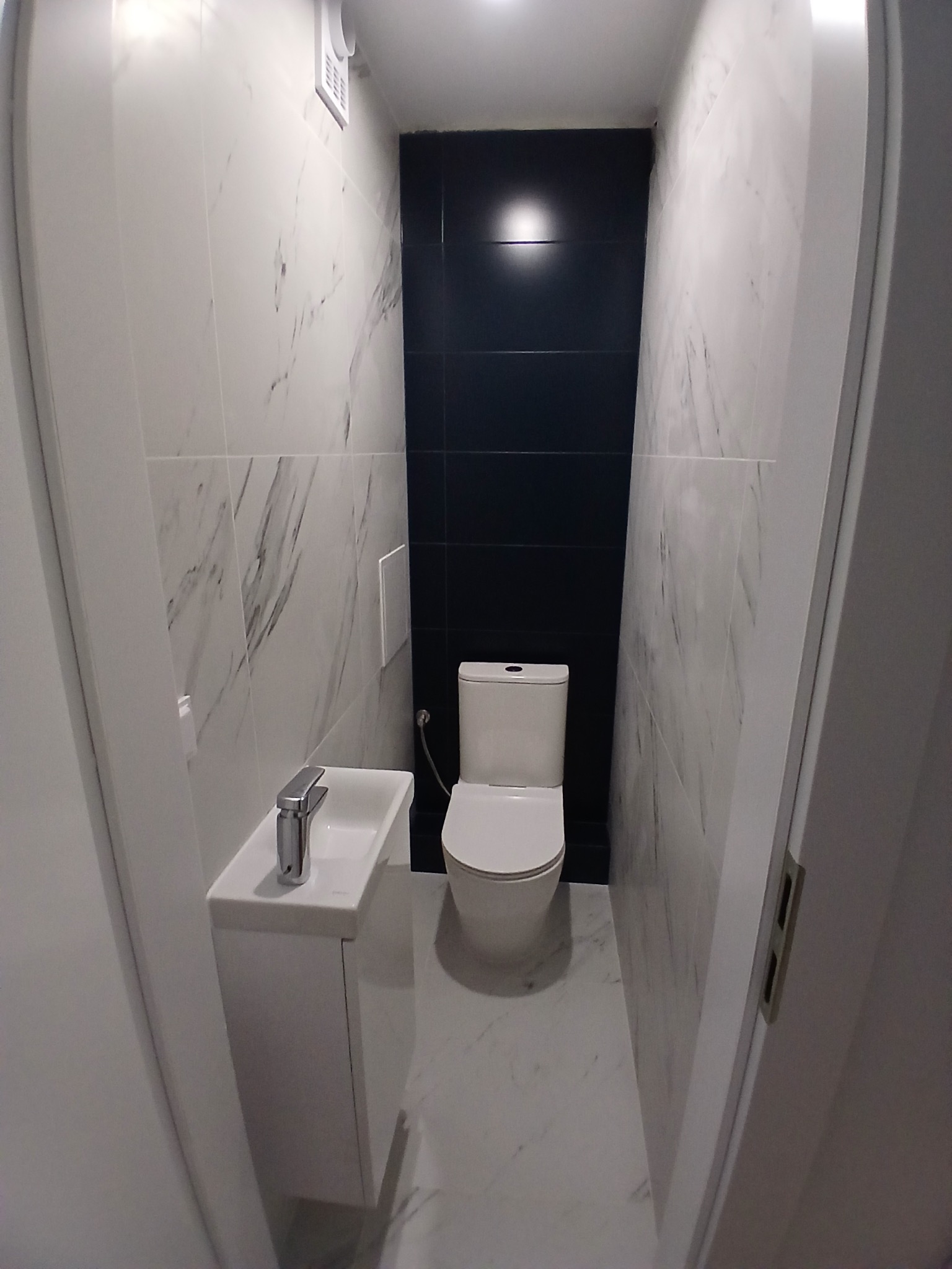 Wąska łazienka z umywalką nablatową i toaletą. Ściany wyłożone białymi płytkami imitującymi marmur, jedna ściana w kolorze granatowym. Minimalistyczny design.