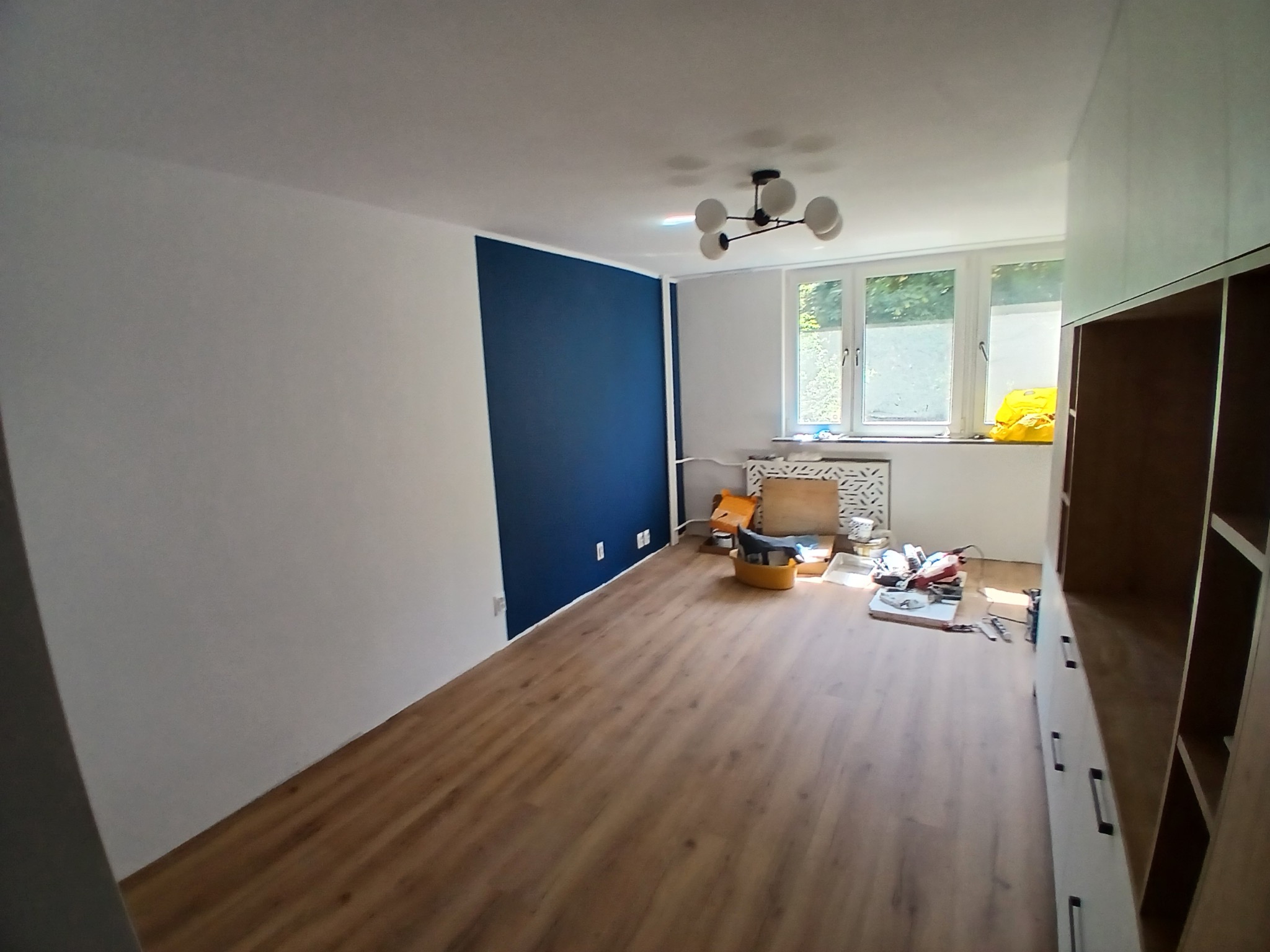 Pokój po częściowym malowaniu ścian na biało i granatowo, z drewnianą podłogą i narzędziami w tle. Widoczna szafa w zabudowie i okno z widokiem na zieleń.