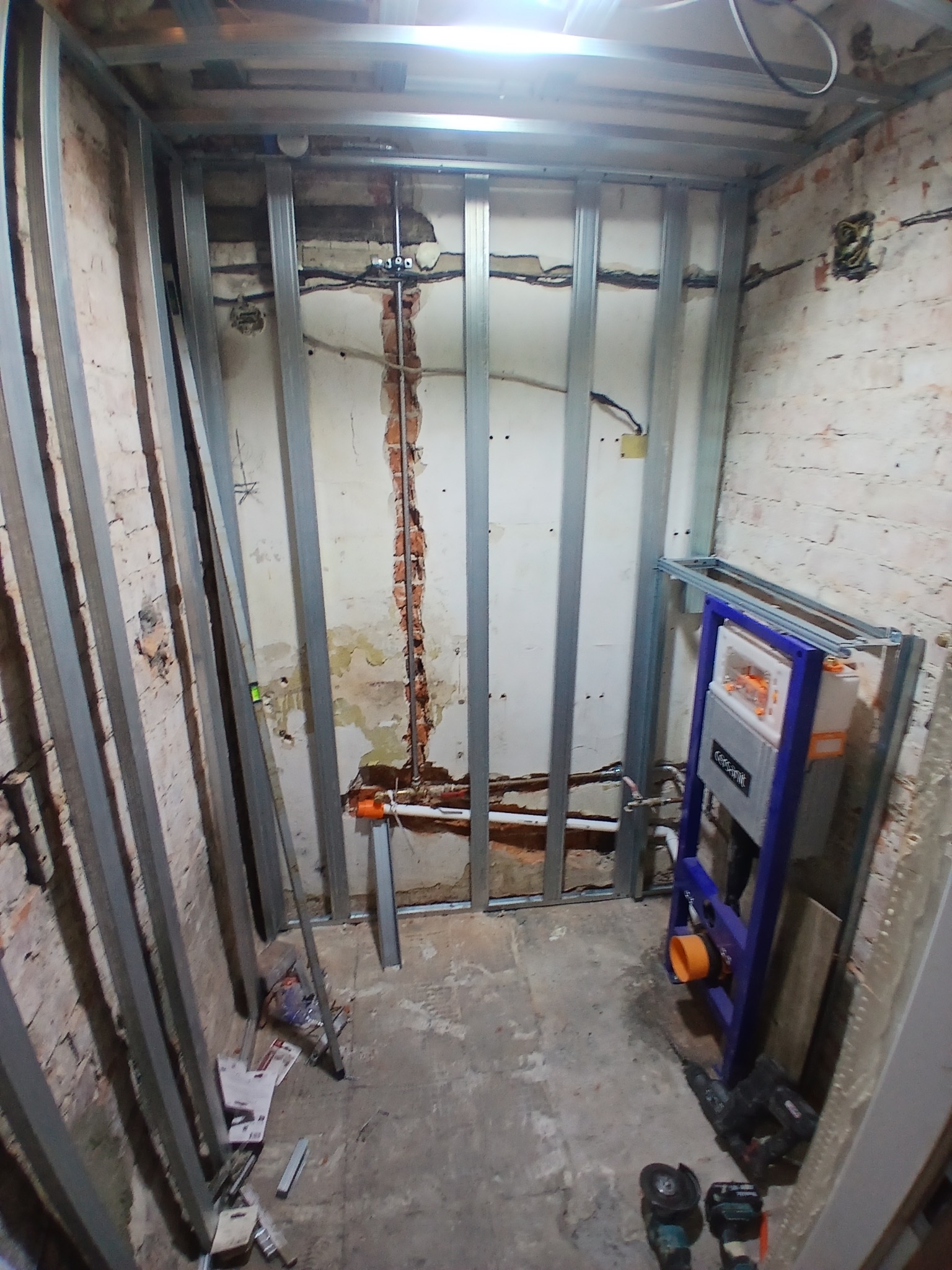 Stelaż metalowy pod zabudowę w surowej łazience z odsłoniętą cegłą. Widoczne narzędzia i instalacja podtynkowa. Etap prac przed wykończeniem.