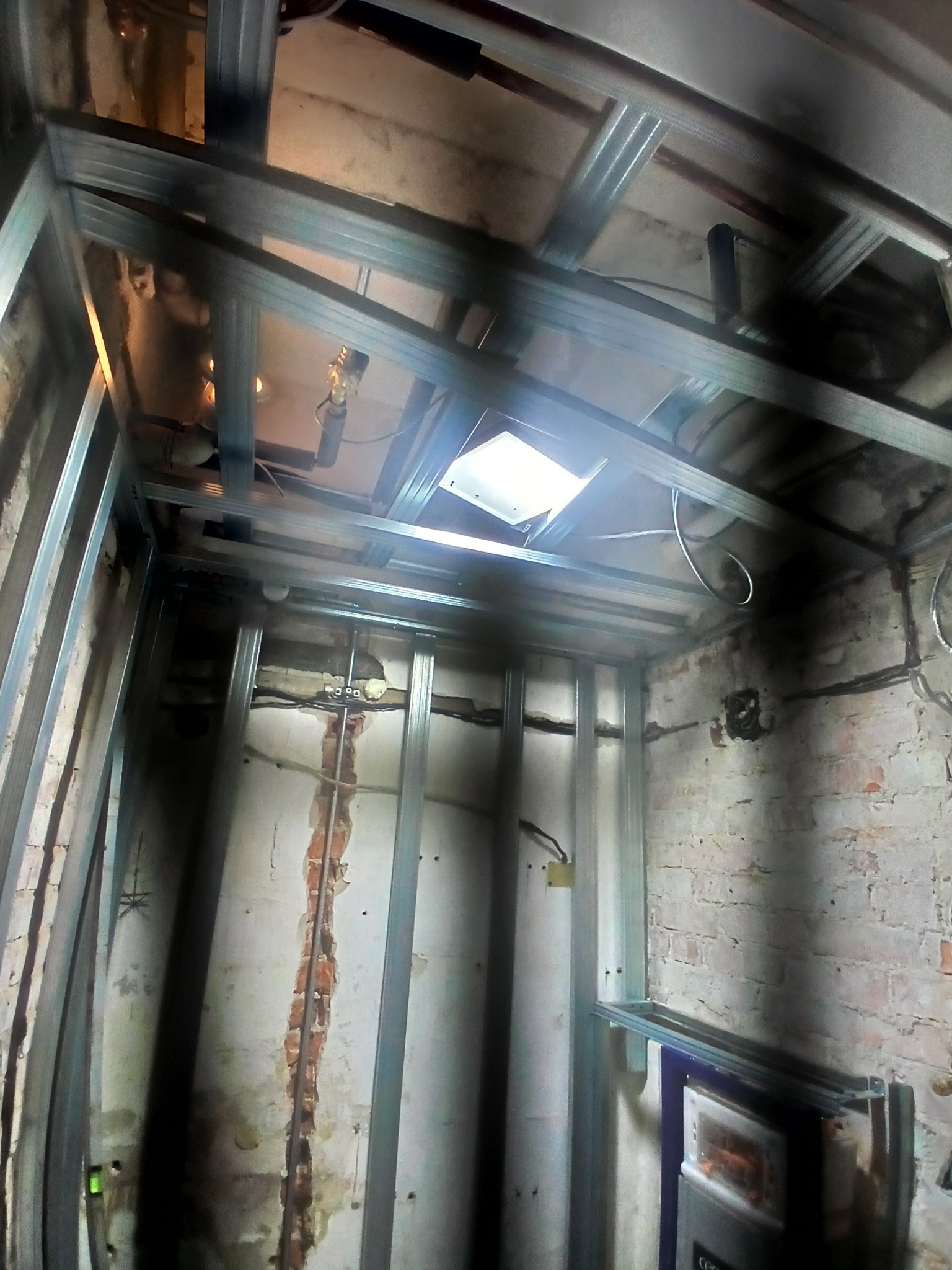 Montaż stelaża z profili metalowych na ścianach i suficie w surowym wnętrzu. Widoczne instalacje i fragmenty starej cegły. Przygotowanie do wykończenia wnętrza.