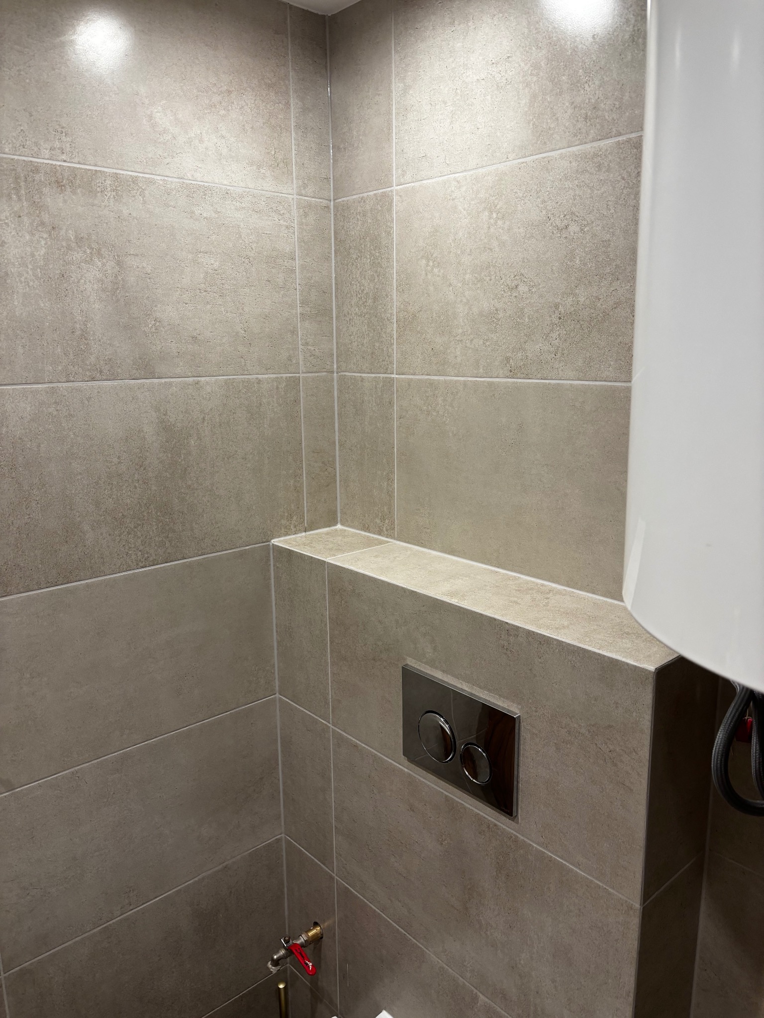 Beżowe, matowe płytki w łazience, ułożone równo na ścianach i półce. Widoczny fragment bojlera i chromowany przycisk spłuczki. Minimalistyczny design.