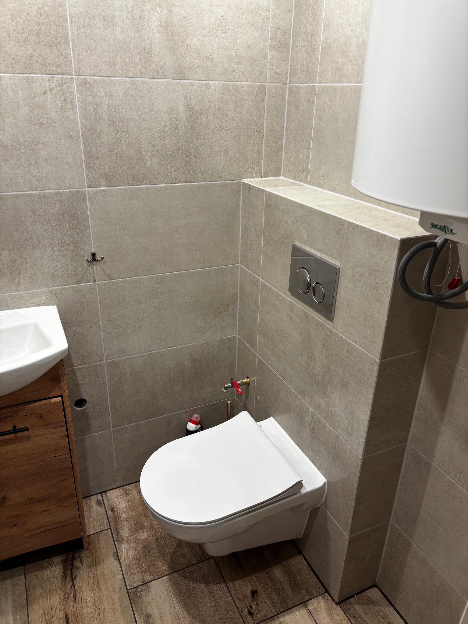 Nowoczesna łazienka z beżowymi płytkami, drewnianą szafką pod umywalką i podwieszaną toaletą. Widoczny bojler i chromowany przycisk spłukiwania.