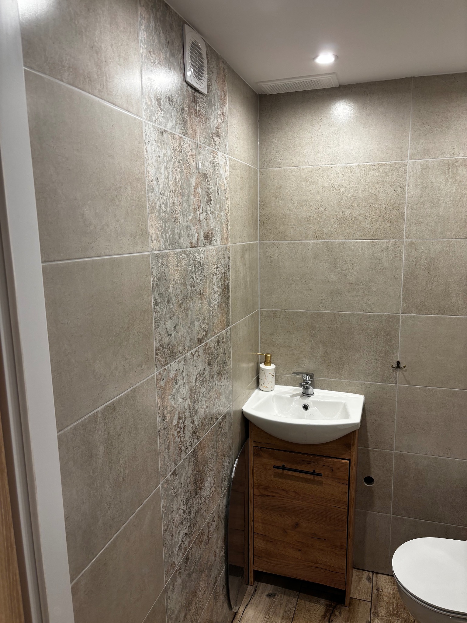 Mała łazienka z beżowymi płytkami imitującymi kamień, drewnianą szafką pod umywalką i toaletą. Widoczny wentylator i oświetlenie sufitowe.