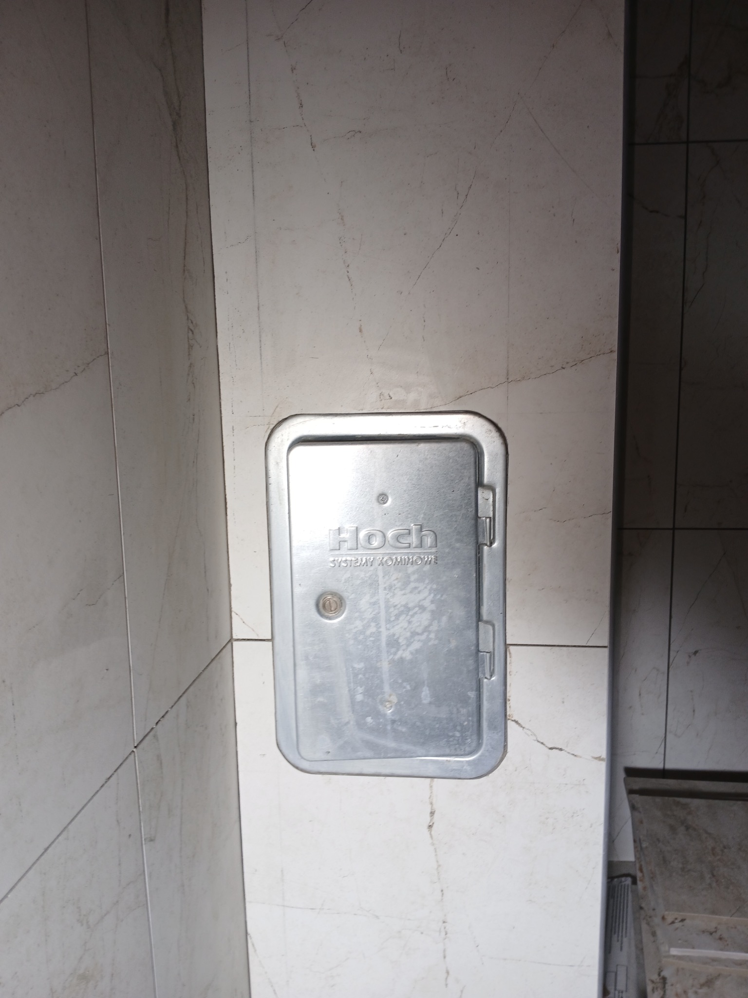 Szara klapka rewizyjna z zamkiem, wkomponowana w marmurowe płytki ścienne. Widoczne fugi i delikatny wzór marmuru. Ujęcie z bliska, skupione na detalu.