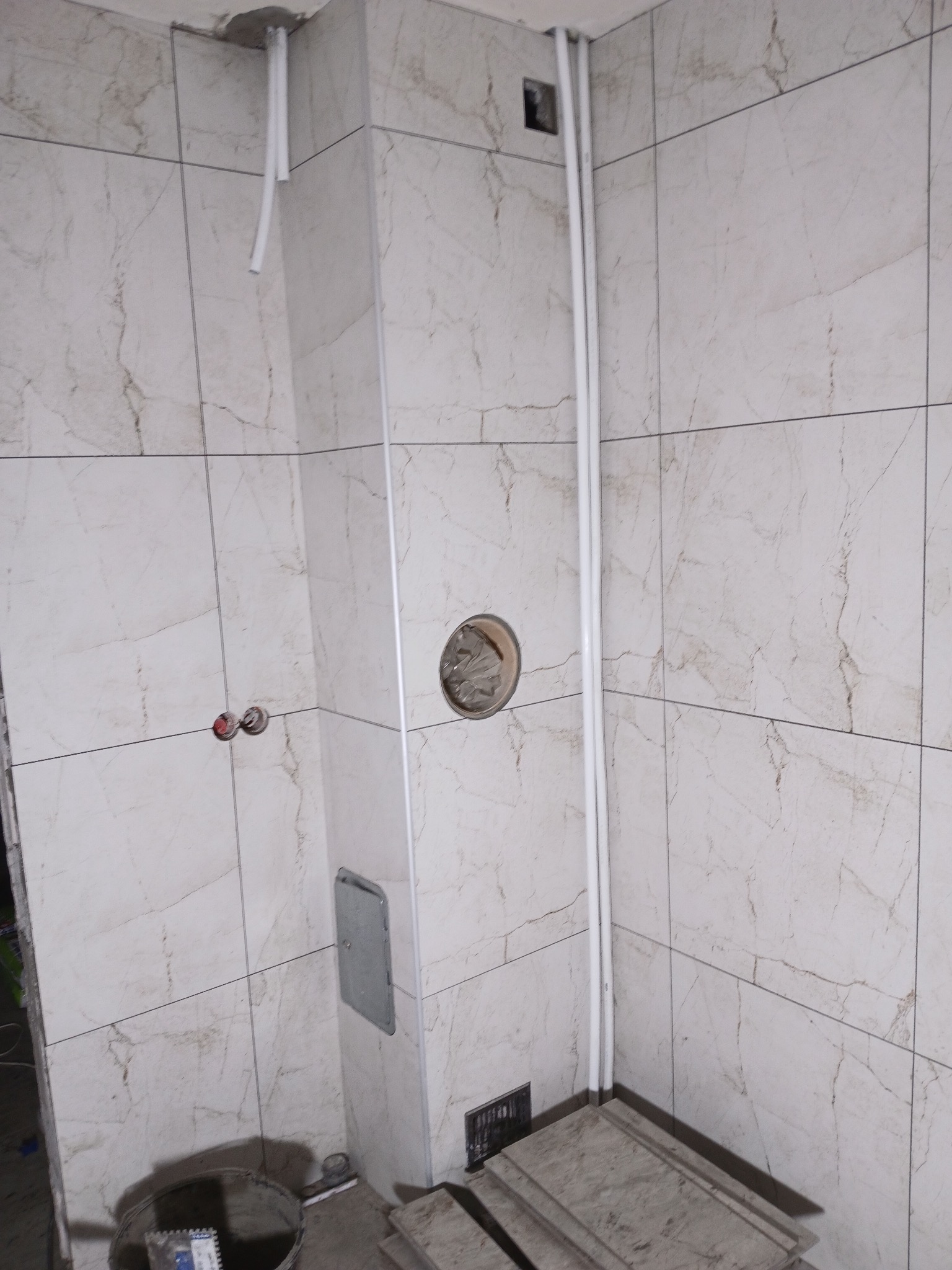 Wykończona łazienka z marmurowymi płytkami, widoczne rury i otwór na baterię. Częściowo widoczne narzędzia i wiadro. Minimalistyczny design z elementami instalacji.