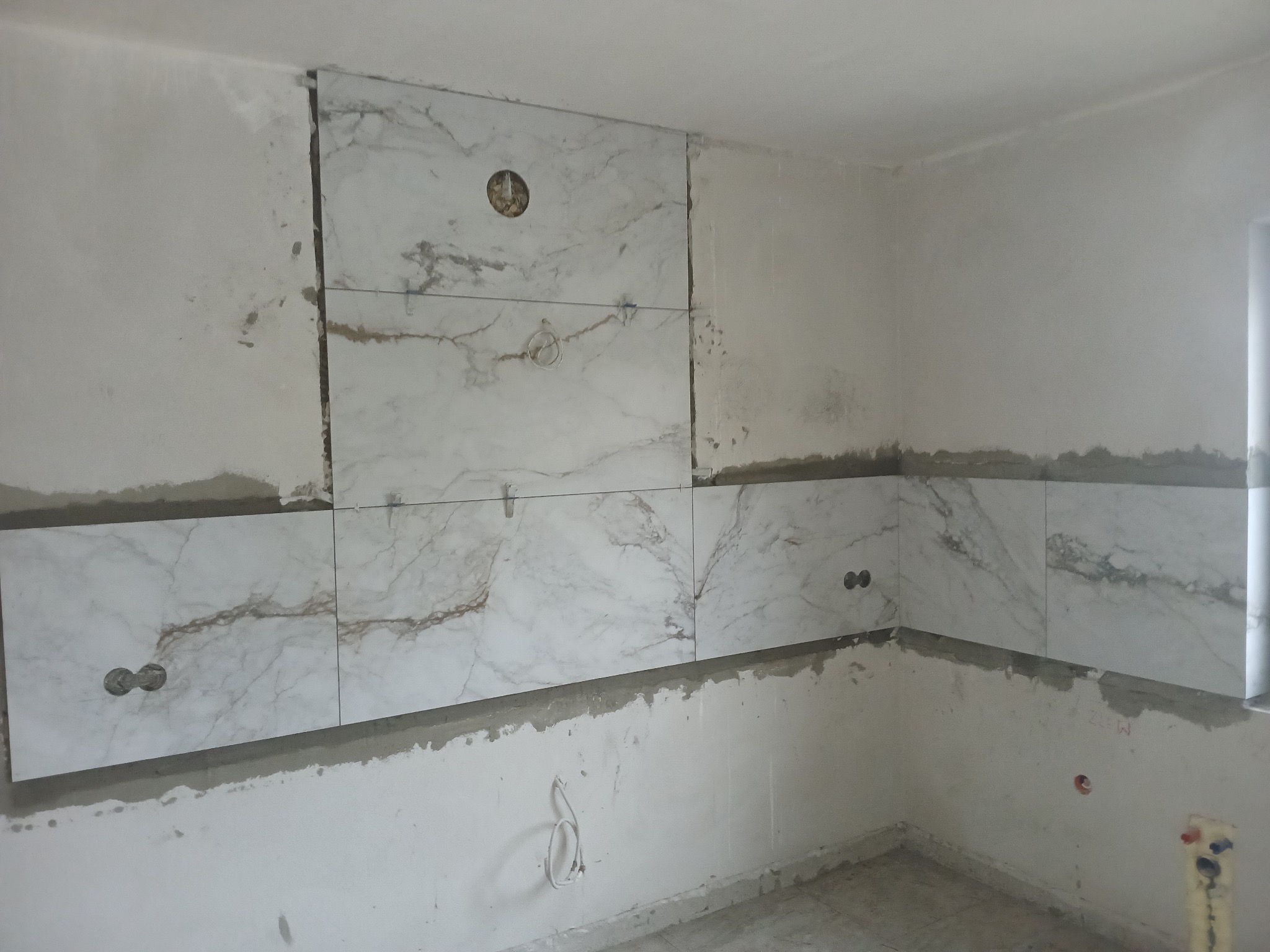 Układanie marmurowych płytek na ścianie w kuchni, widoczne otwory na gniazdka i rury, etap prac wykończeniowych, surowy stan ścian, szare fugi.