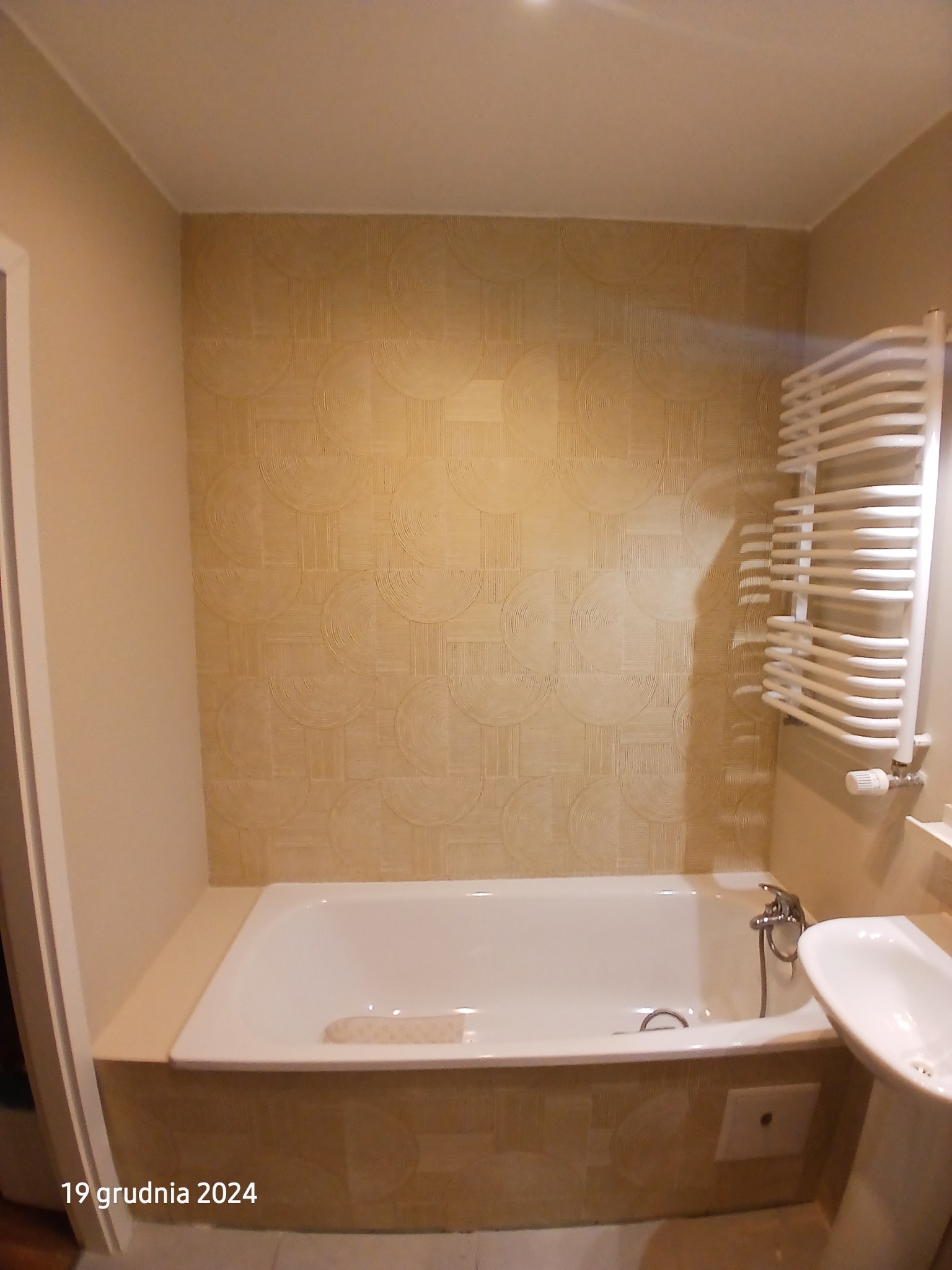 Wykończona łazienka w Pęgowie: biała wanna z baterią, umywalka na postumencie i grzejnik drabinkowy na ścianie z wzorzystą tapetą w ciepłych odcieniach.